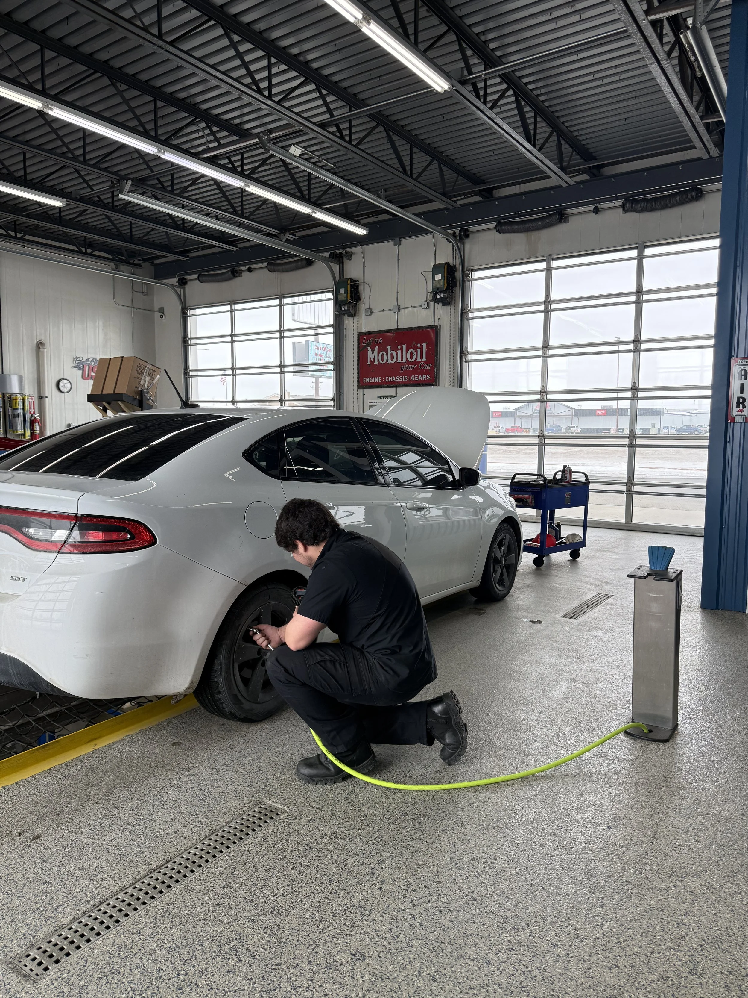 A mechanic kneeling next to a white sedan with the hood open, working on the rear tire in an auto repair shop.