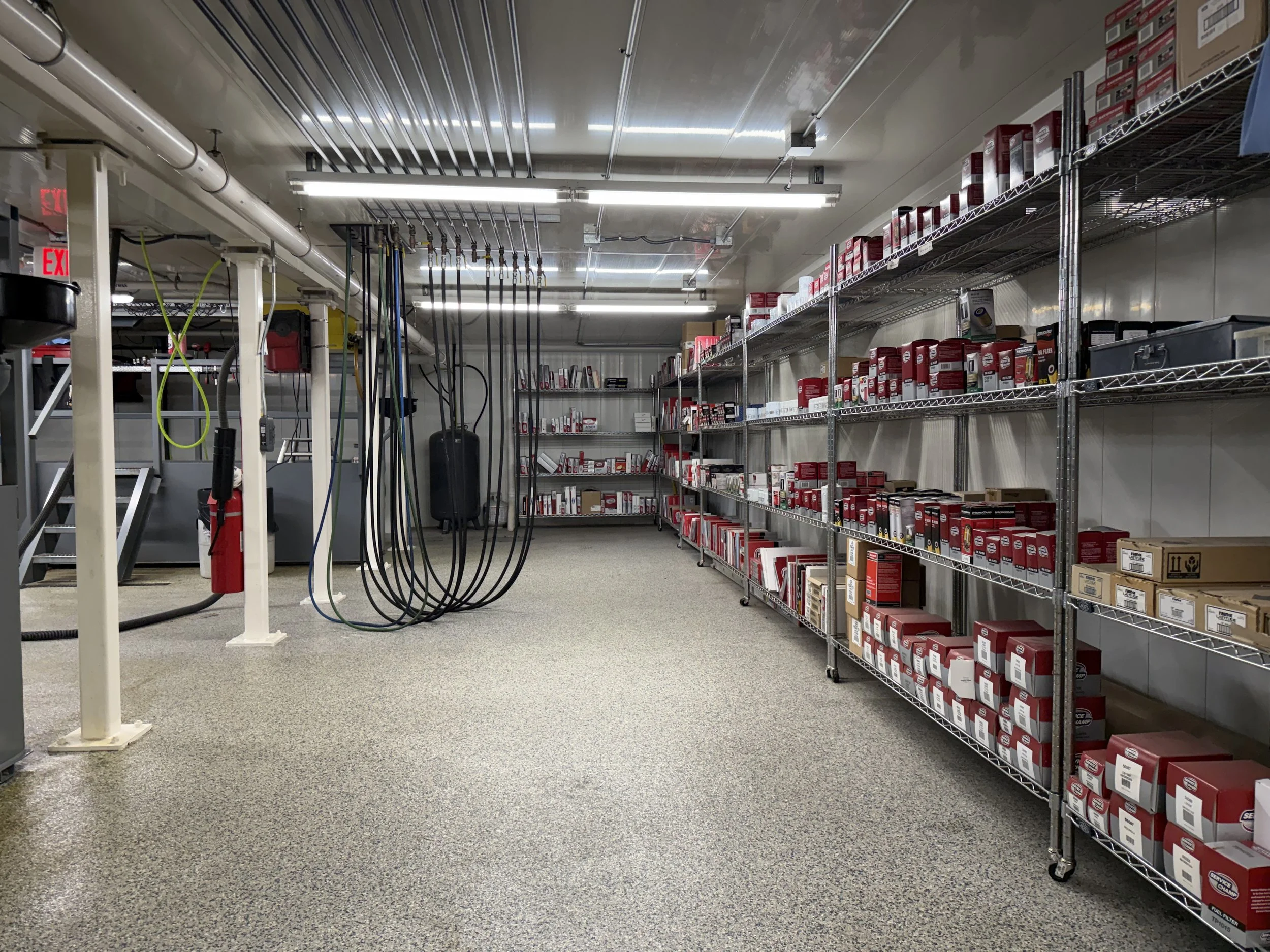 An industrial storage room with metal shelves filled with red and brown boxes, and a white metal rack with tools and equipment hanging from hooks. The floor is carpeted, and the ceiling has bright fluorescent lights. There are hoses hanging on a white support beam, and a fire extinguisher near the support.