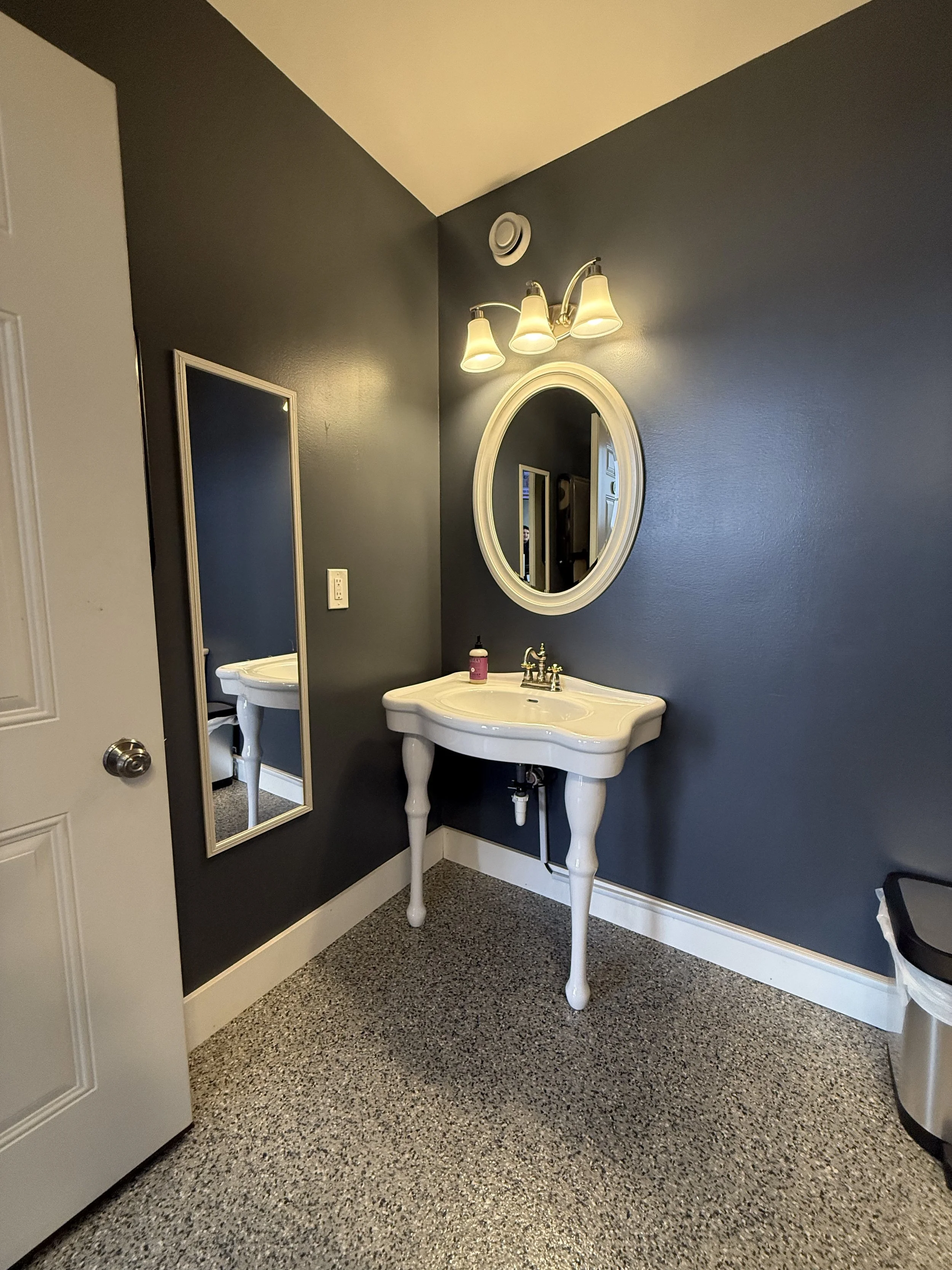A bathroom with a white vintage-style sink, a round mirror above it, a wall-mounted light fixture with three bulbs, a full-length mirror on the dark gray wall, and a trash can on the right.