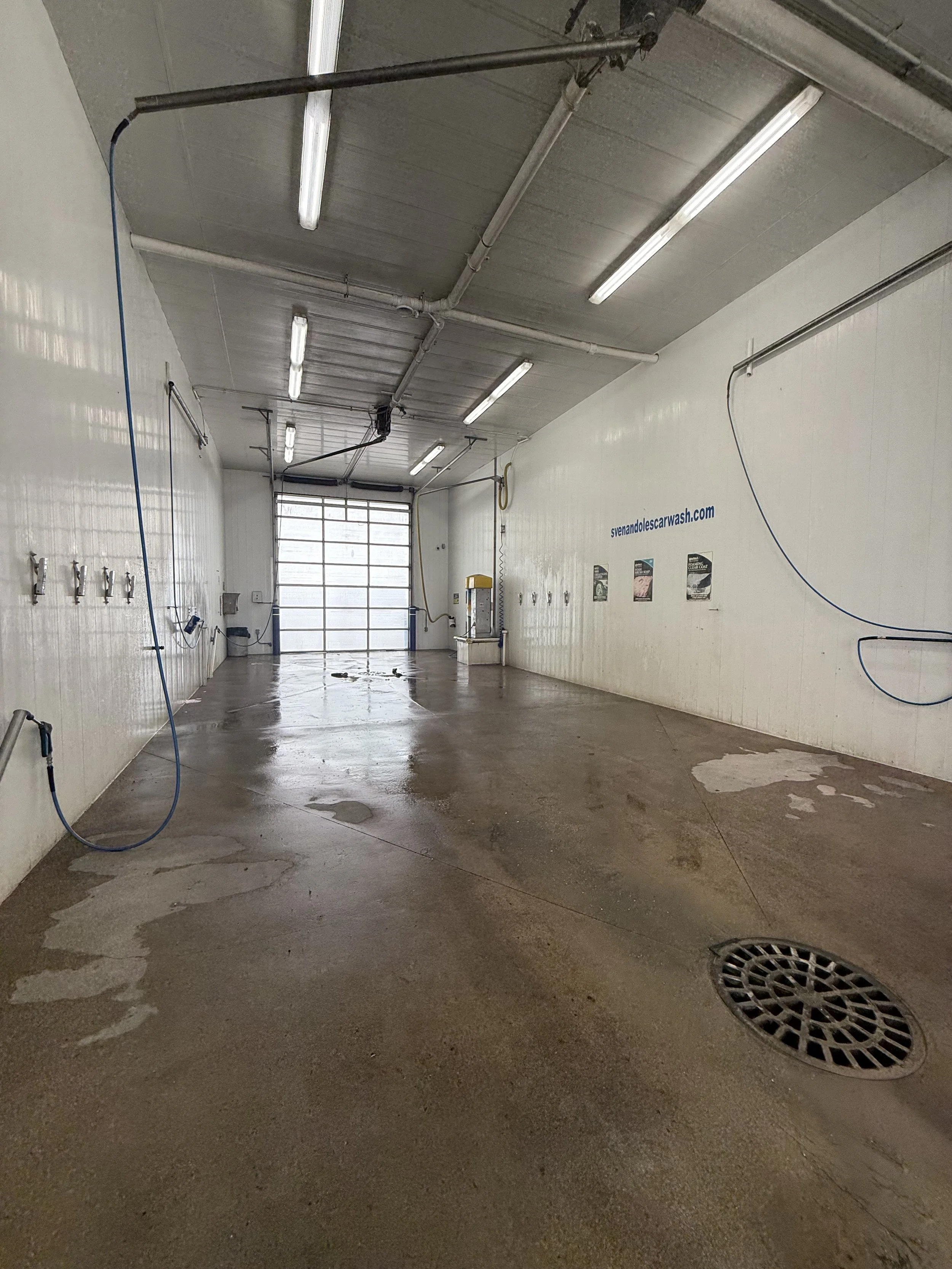 An empty car wash bay with wet concrete floor, water hoses hanging on the walls, bright fluorescent ceiling lights, a large roller door at the back, and posters on the wall.