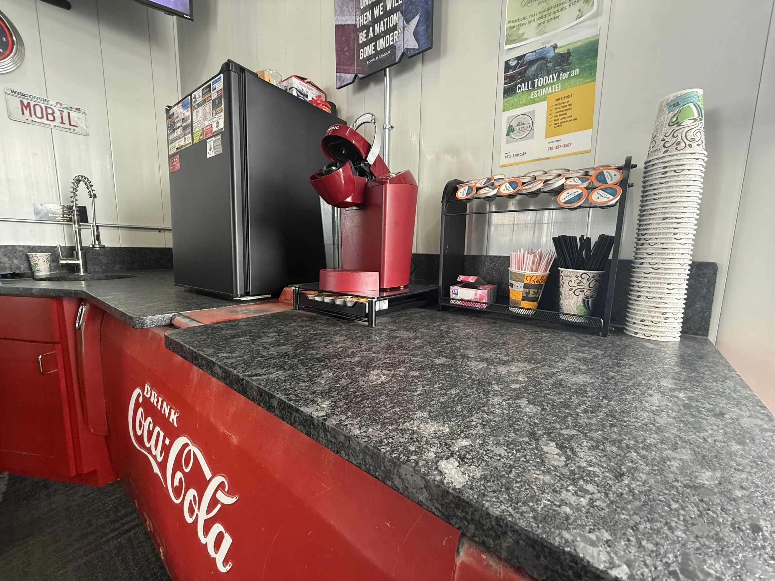Coffee station with a red coffee maker, disposable cups, stir sticks, and a small refrigerator on a black granite counter against a wall with advertisements.