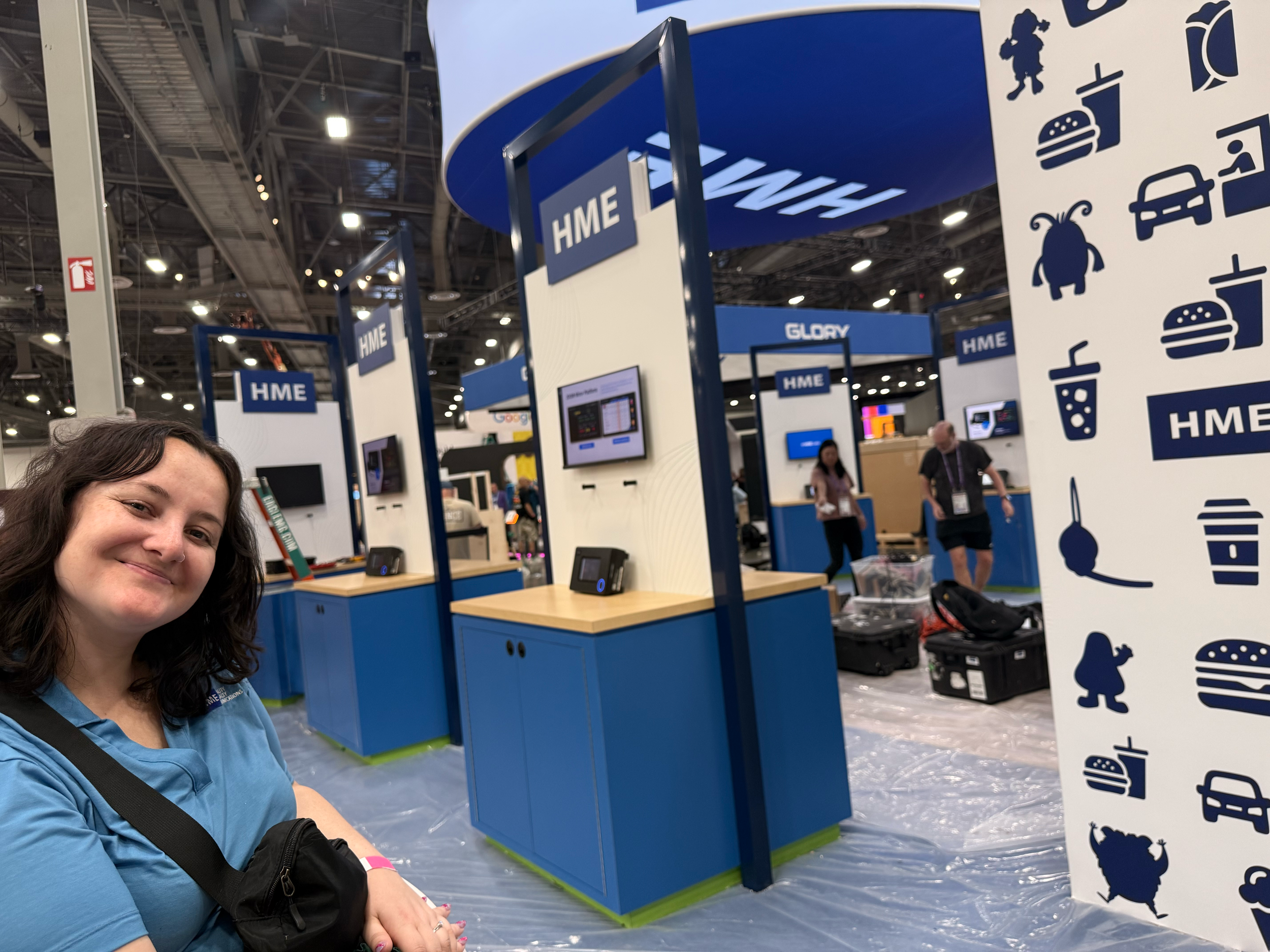 Jordan Cornejo in a blue shirt setting up the the HME booth at McDonald's Worldwide Convention. The background shows blue and white booth displays with signs labeled "HME" and various icons of food, animals, and other items.