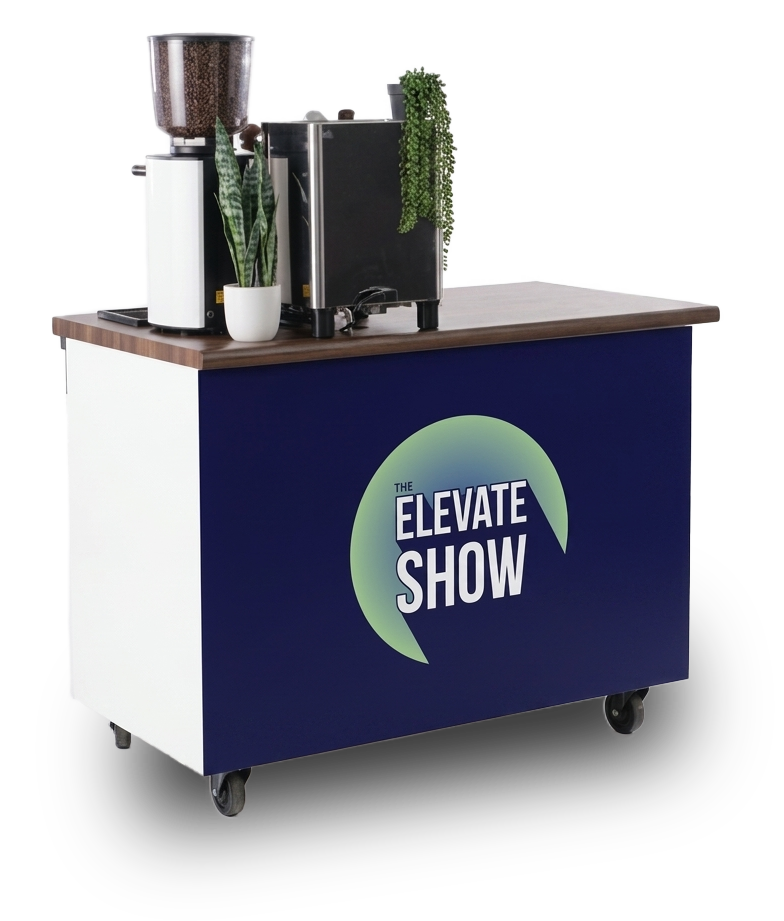 A mobile counter with a sign reading 'The Elevate Show' on the front, topped with a coffee machine, a blender, and potted plants.