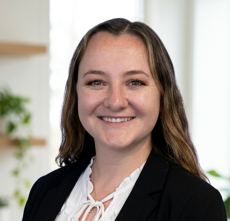 A smiling woman with shoulder-length wavy brown hair, wearing a white blouse and black blazer, standing indoors with plants and a bookshelf in the background.