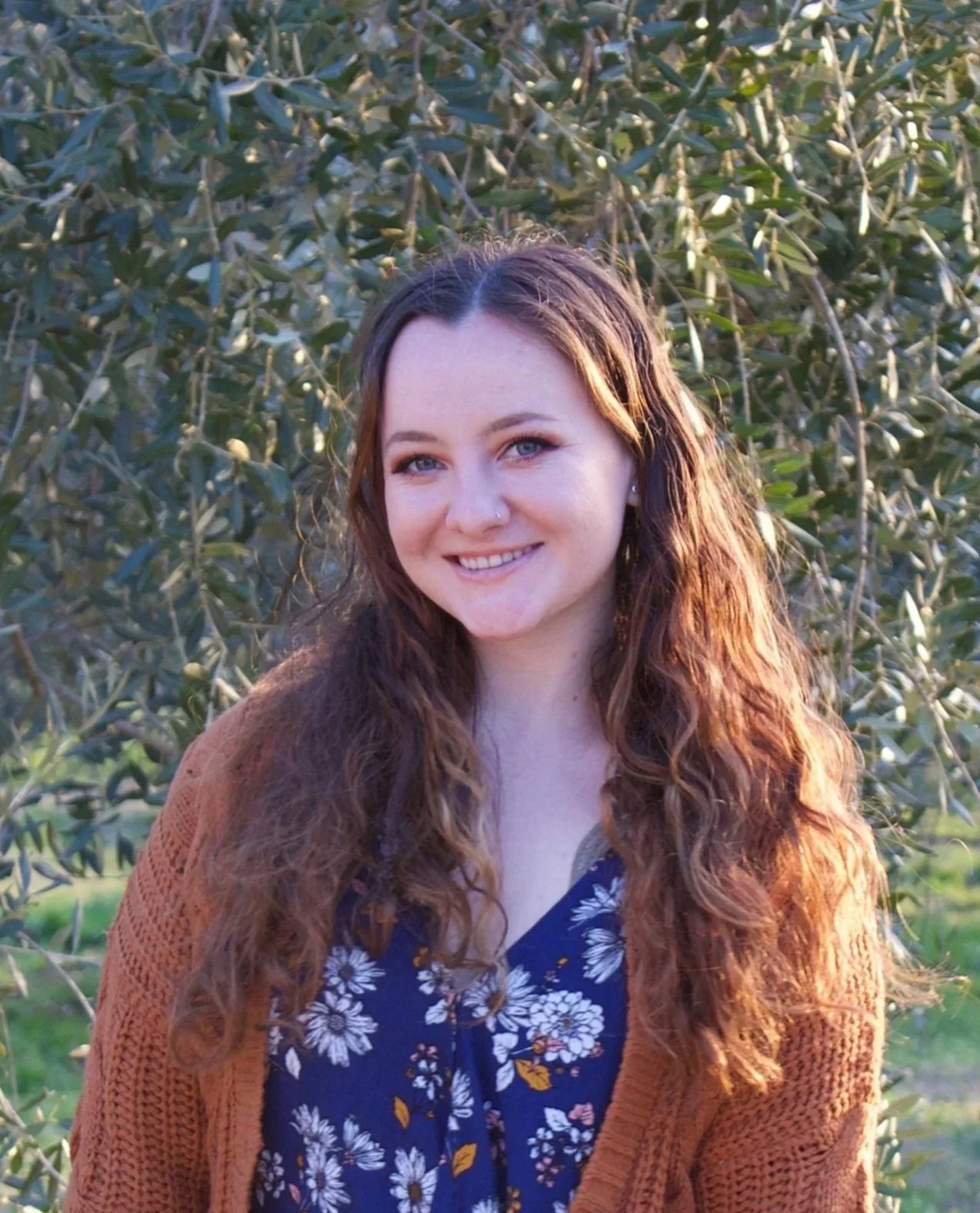 A woman with long, wavy brown hair and blue eyes smiling outdoors in front of green foliage, wearing a floral dress and a brown cardigan.