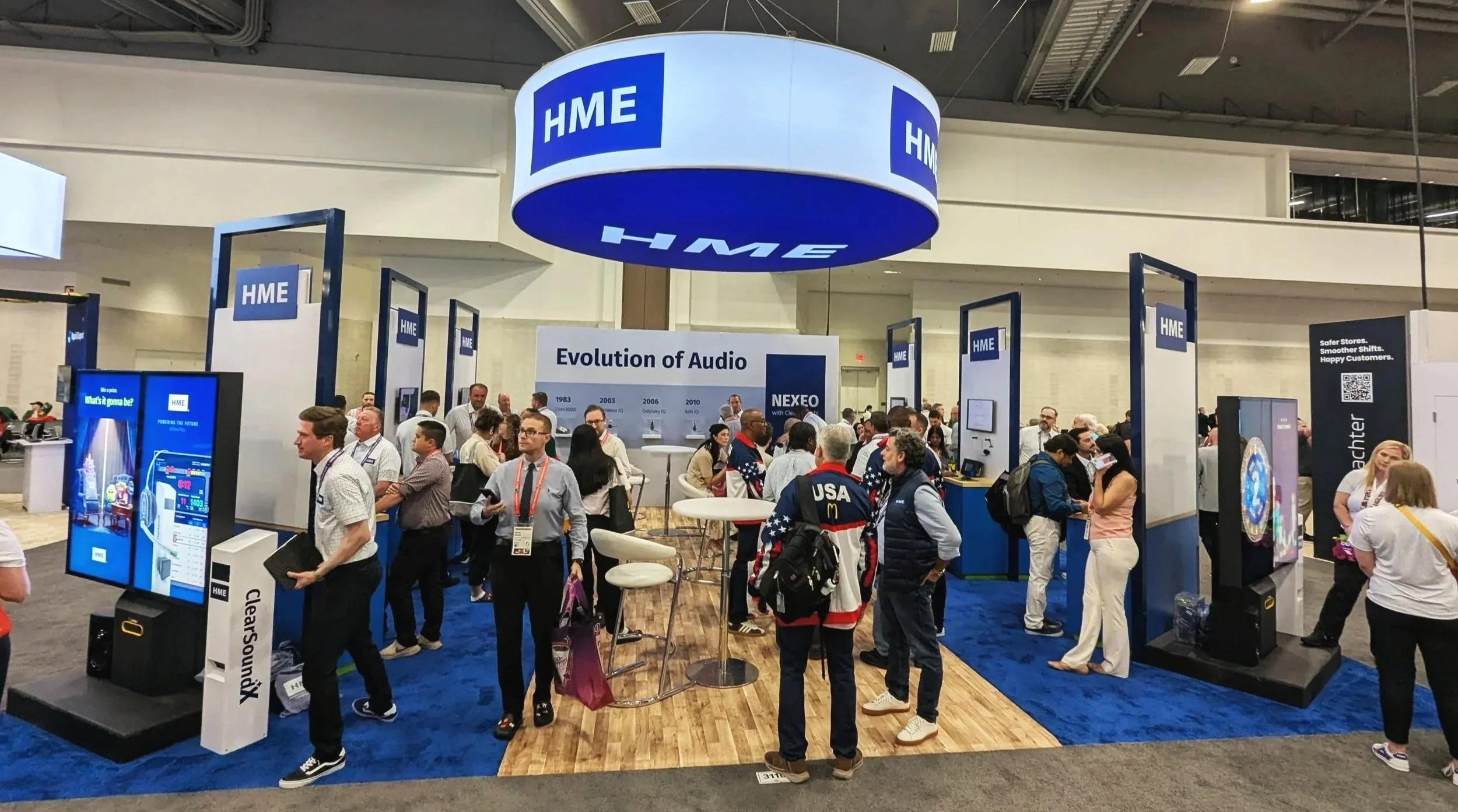 Trade show booth for HME, featuring a large hanging sign with the HME logo, informational displays about the evolution of audio technology, and a crowd of attendees engaging with the exhibits and representatives.