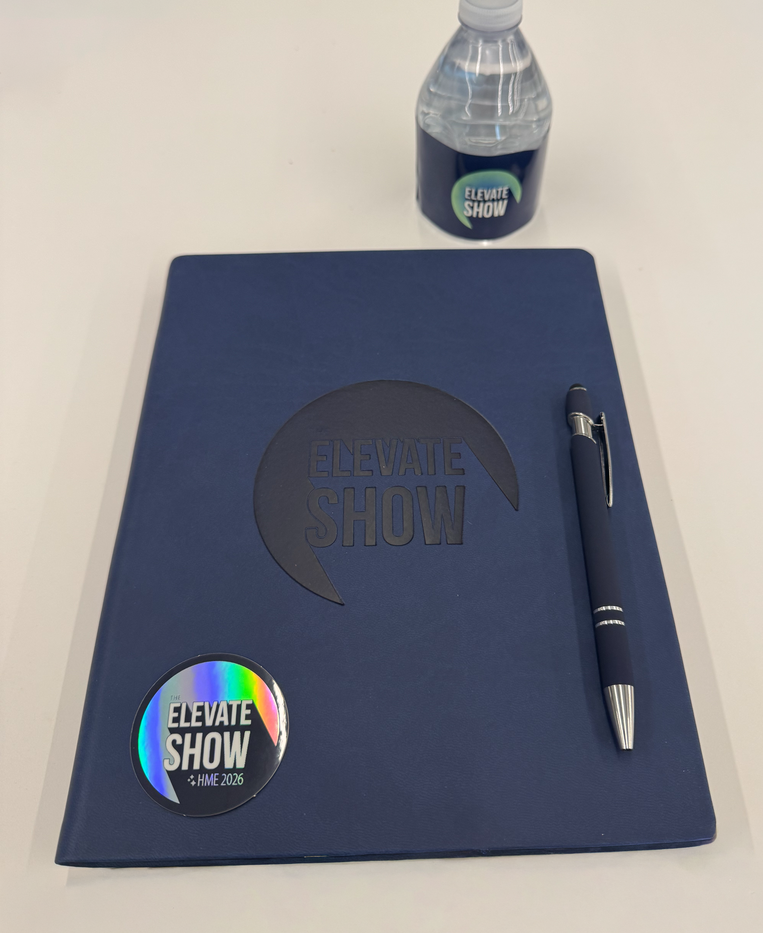 A blue notebook with the logo 'Elevate Show' on the cover, a silver pen beside it, a bottled water labeled 'Elevate Show,' and a 'Elevate Show' sticker on the notebook.