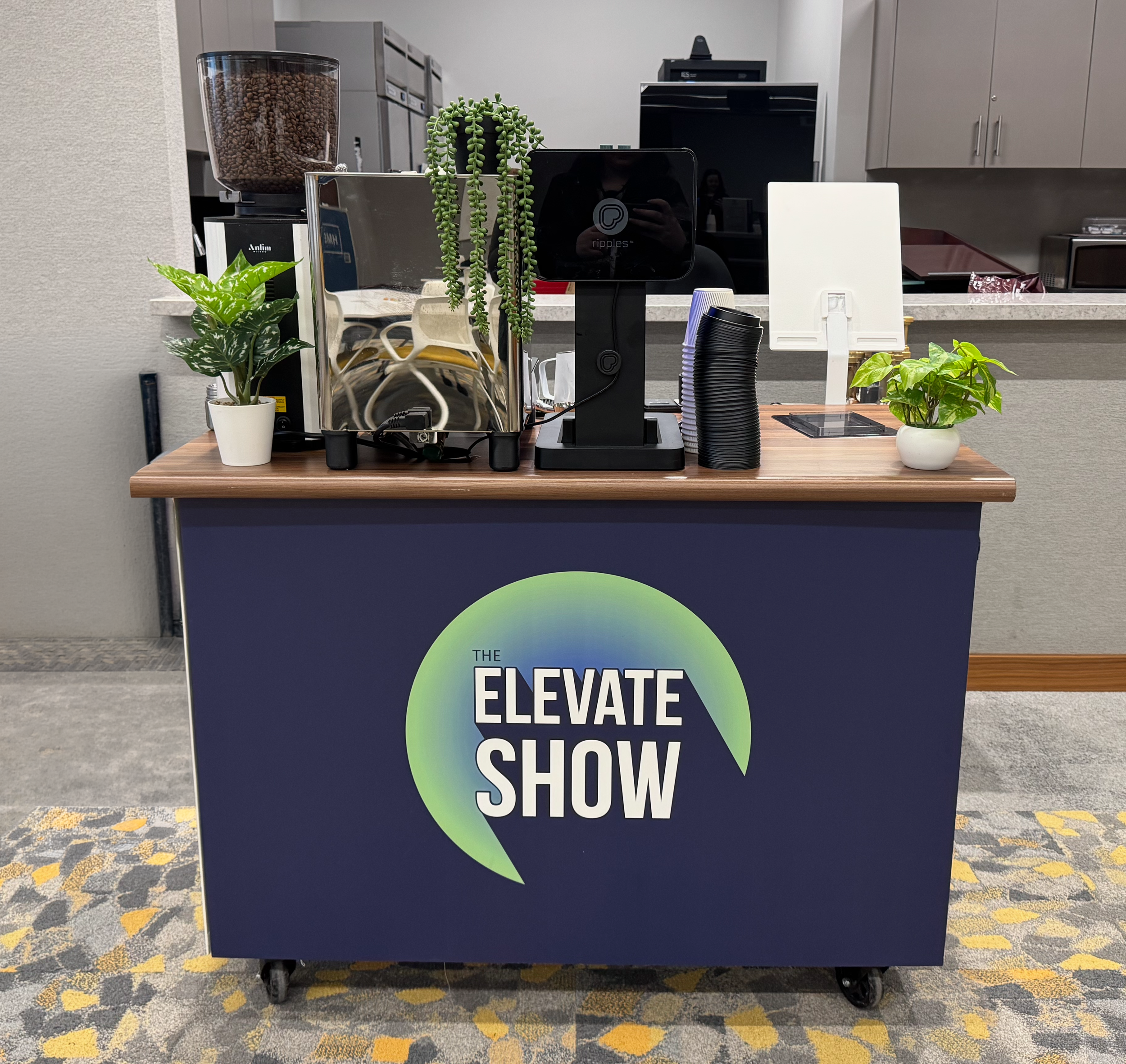 A counter with a sign reading 'The Elevate Show' on the front. The surface holds a coffee bean grinder, a small potted plant, a digital device, a stack of black cups, and another small plant in a white pot. The background includes a kitchen area with cabinets and appliances.