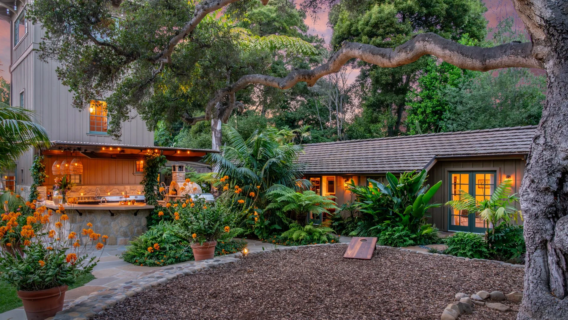 evening view of Sea Ranch outdoor kitchen with thatched awning, illuminated hanging lanterns, tea candles, stone counter and pizza oven next to pool cottage and pickle ball court under an ancient oak tree