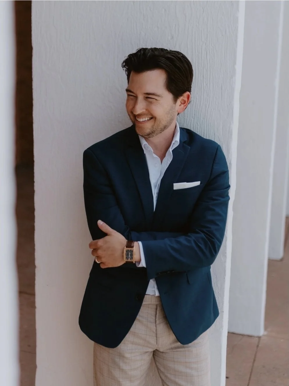 Man in a dark blazer smiling while leaning against a white column.