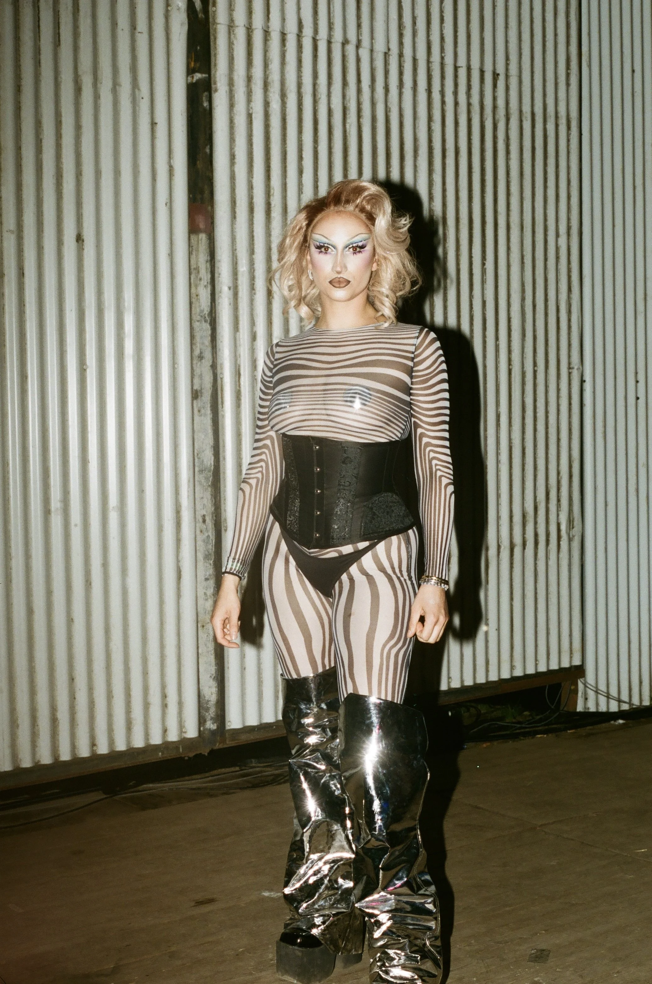 A person with elaborate makeup and blonde curly hair, wearing a sheer striped top, a black corset, matching striped pants, and shiny silver thigh-high boots, standing against a corrugated metal wall.