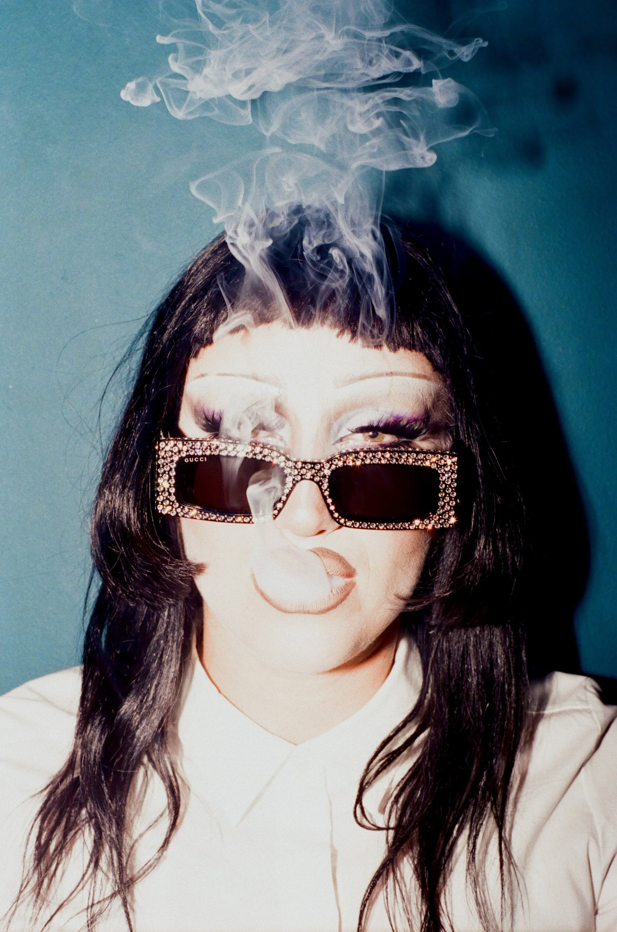 Person with long dark hair wearing large, rhinestone-studded sunglasses, blowing smoke against a teal background.