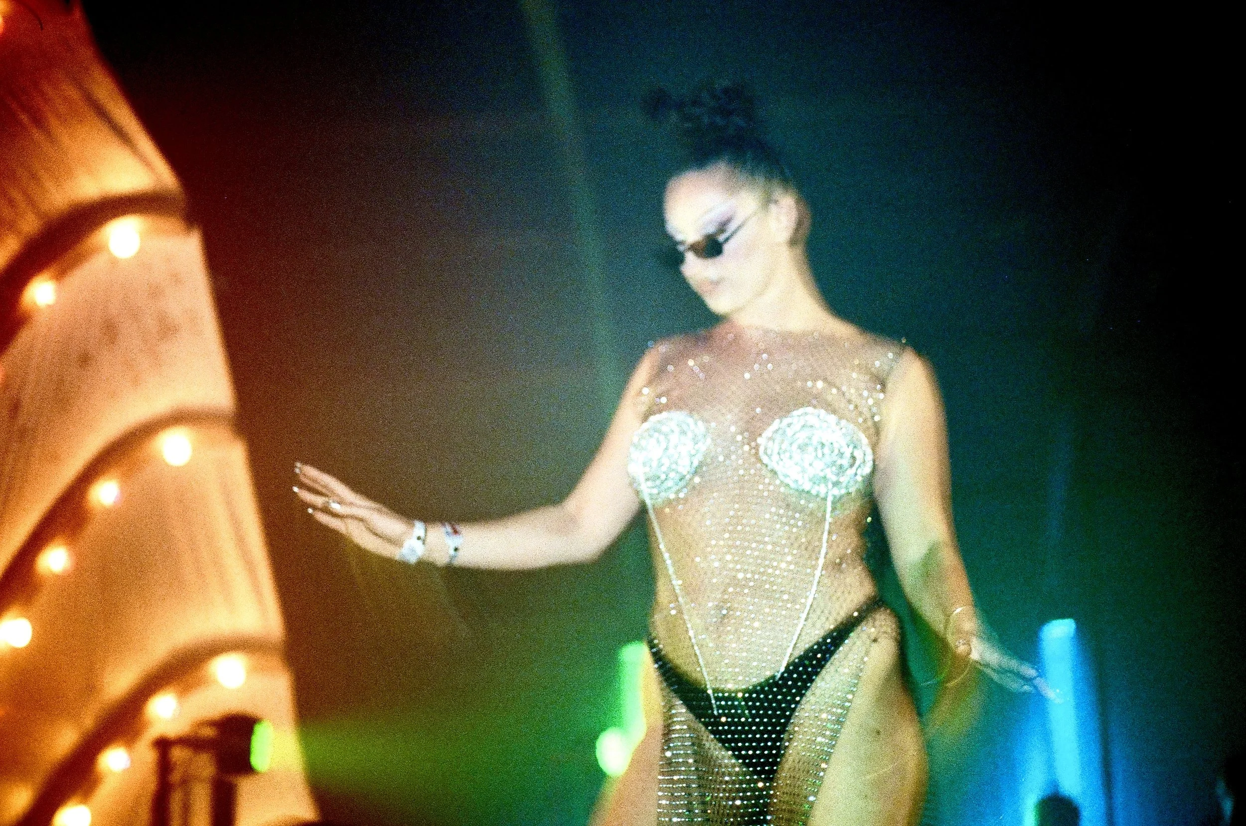 A person dressed in a sparkly, revealing outfit with glasses and a high bun, dancing on a stage with colorful lights.