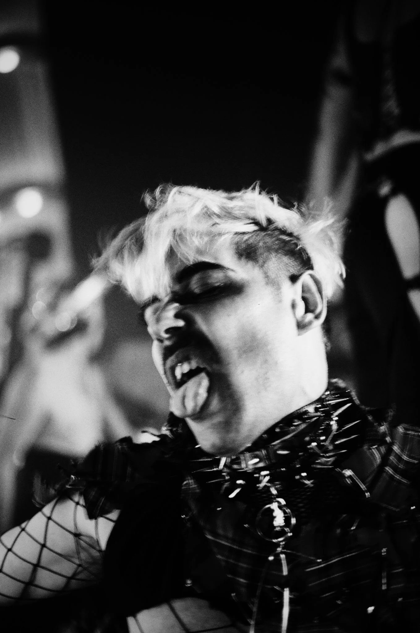 A black and white photo of a person with light-colored, messy hair, sticking out their tongue and making a playful, rebellious face. They are wearing a plaid outfit with spikes and fishnet sleeves.