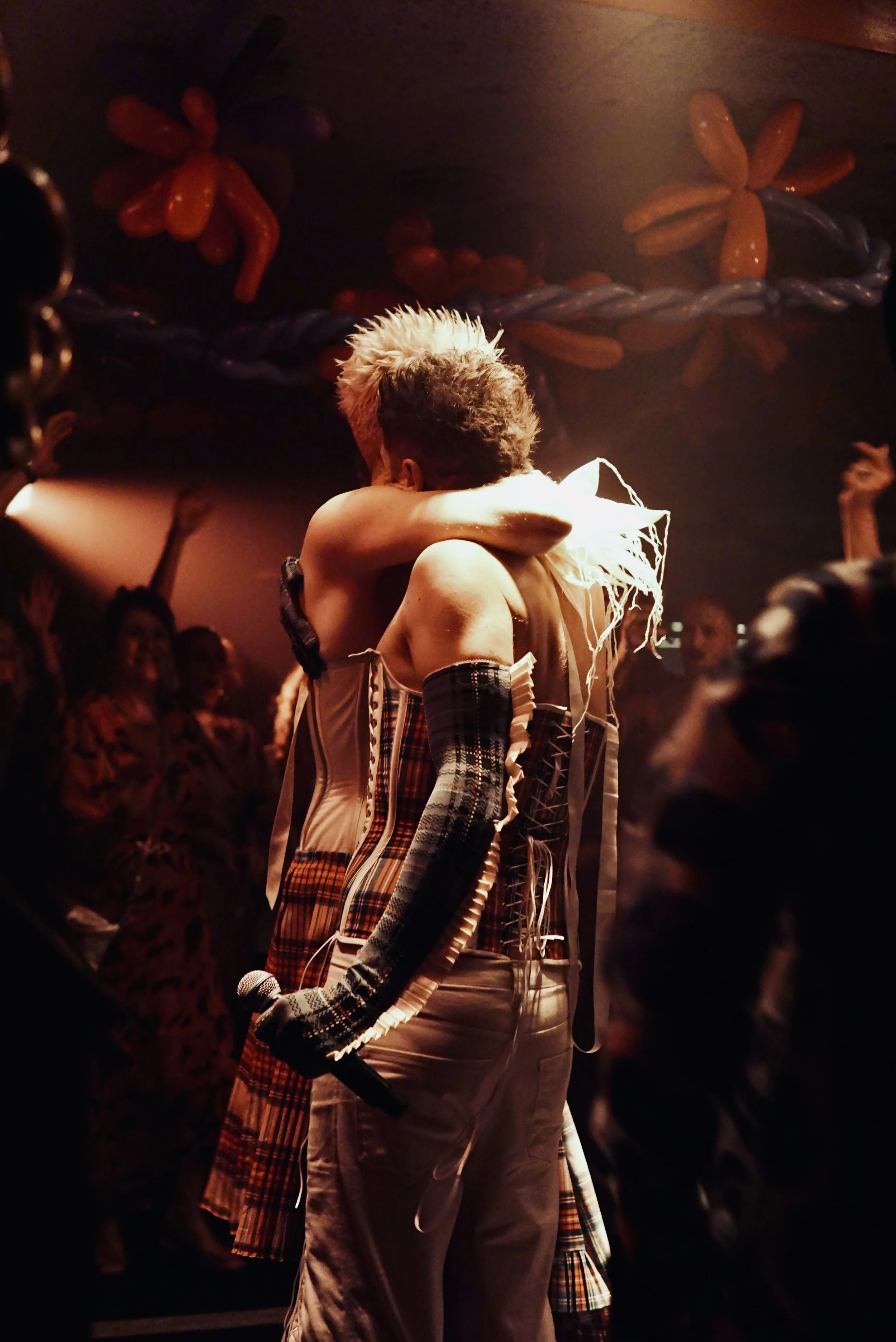 Two performers on stage embracing, one holding a microphone. They are dressed in eclectic, plaid outfits, and there are lively, excited audience members in the background.