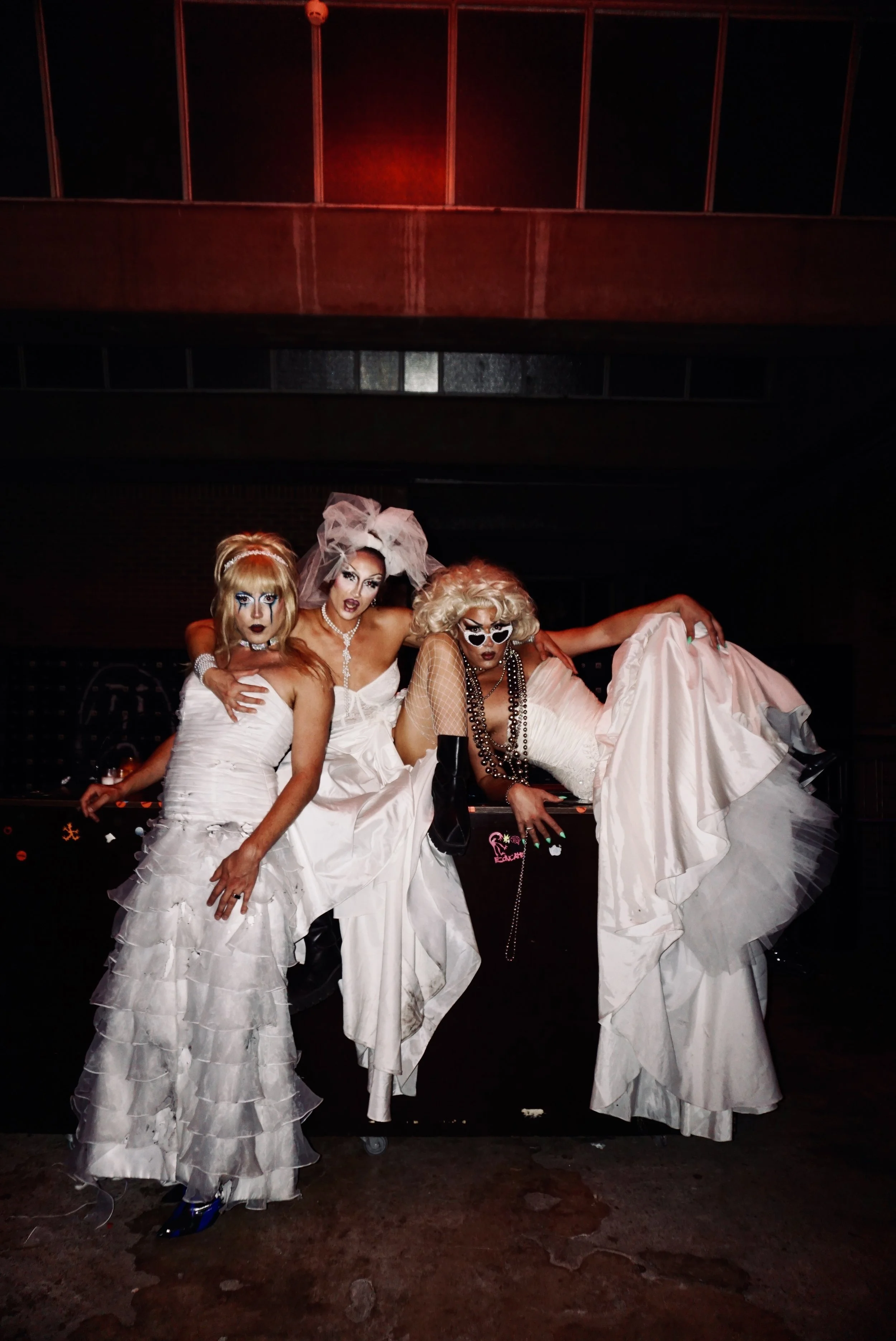 Three drag queens dressed in elaborate white and black costumes posing together in a dark club setting.