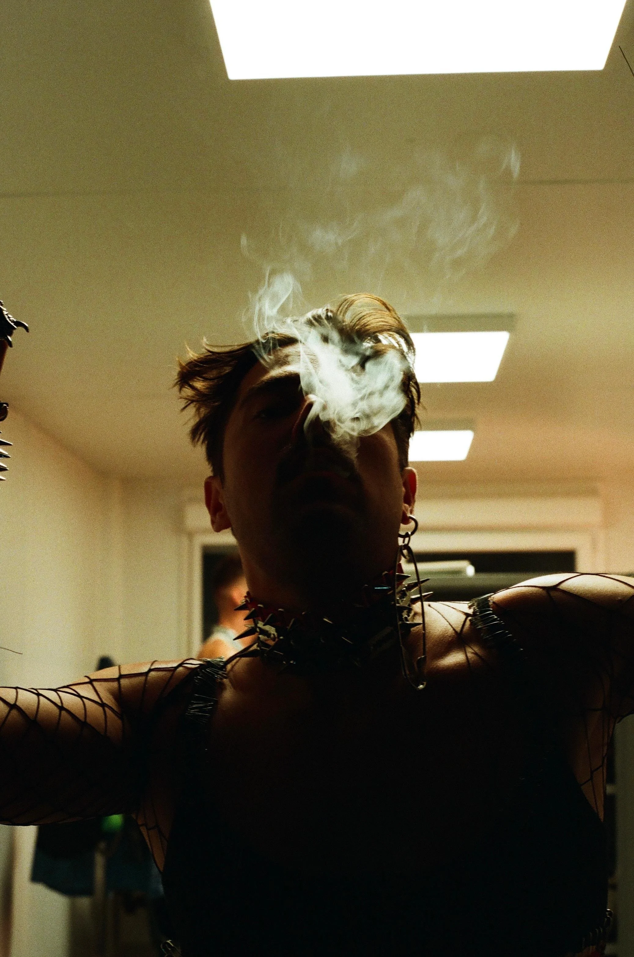 Person with spiked jewelry and fishnet clothing, smoking in a dimly lit room.