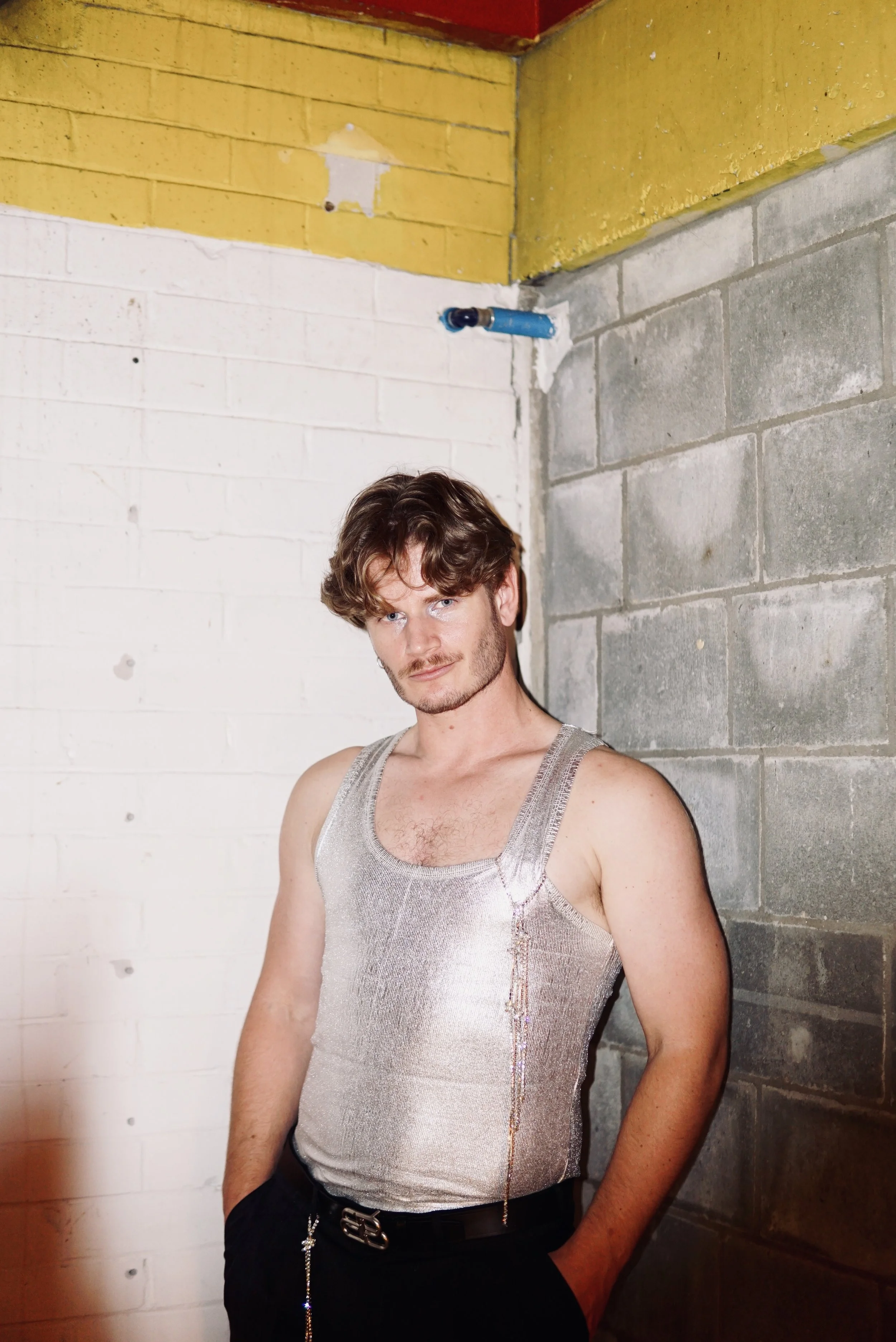 A young man with brown hair and a beard, wearing a shiny silver tank top, stands against a corner wall of cinder blocks and painted bricks in an unfinished space.