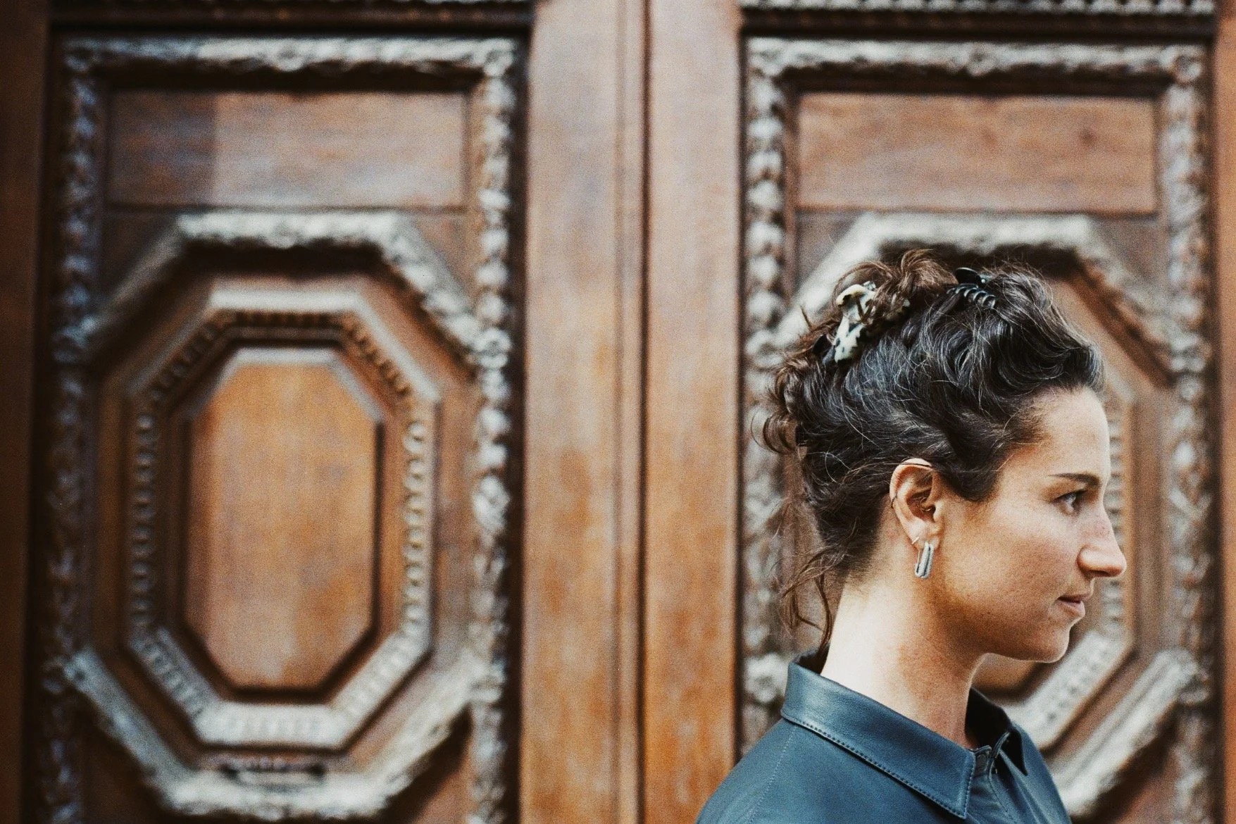 A woman with dark hair styled in an updo with a hair accessory, wearing a blue collared shirt, looking to the right in front of ornate wooden paneling.