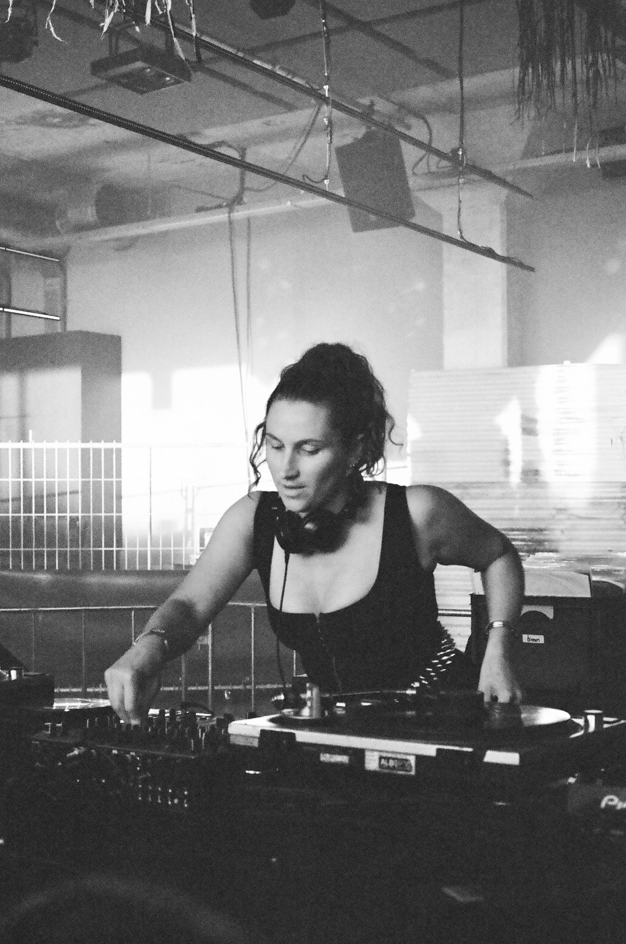 A female DJ with dark curly hair, wearing a sleeveless top, is playing music on turntables in a dimly lit, industrial-style setting.