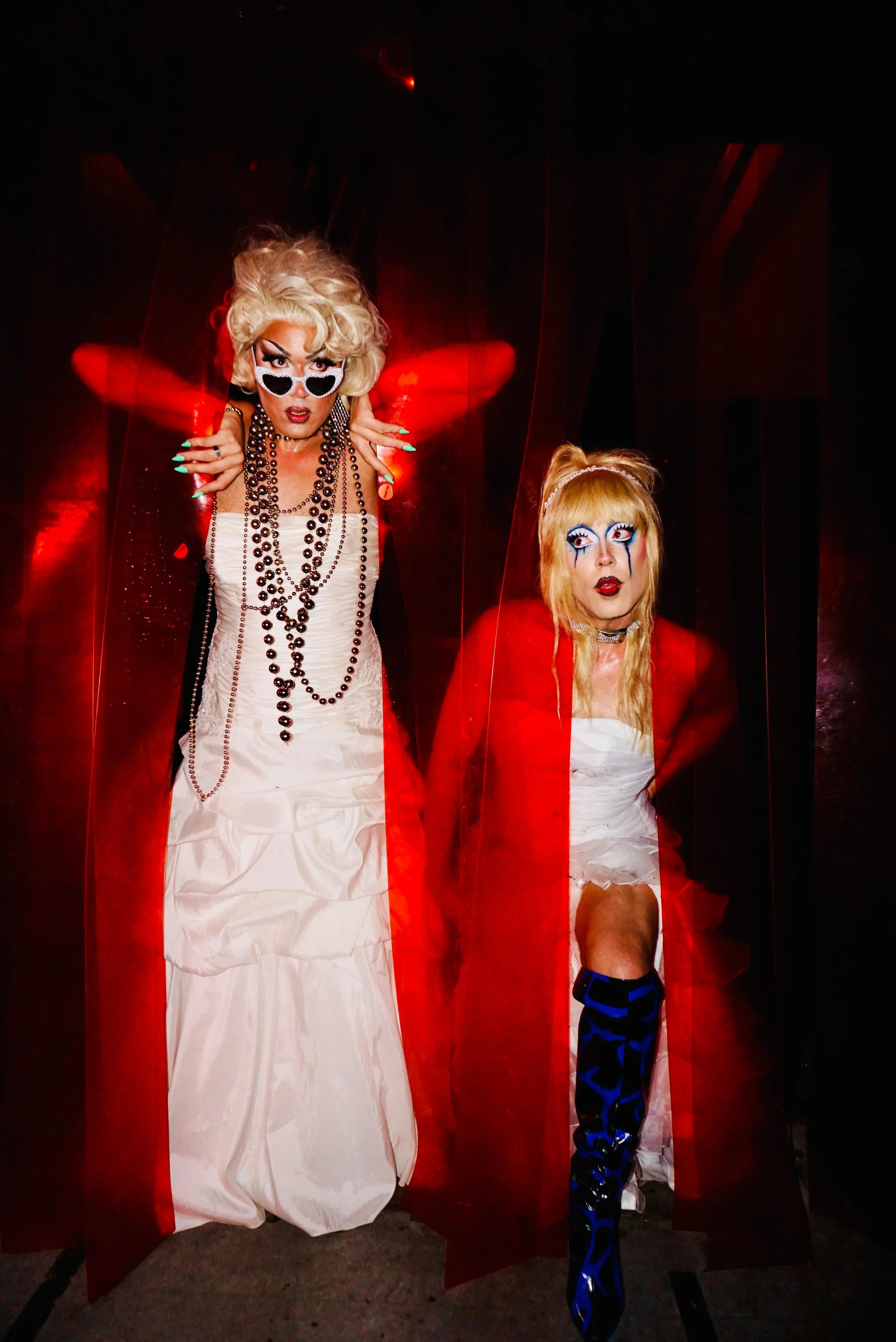Two drag queens with bold makeup and costumes, posing in front of a dark background with red lighting effect.