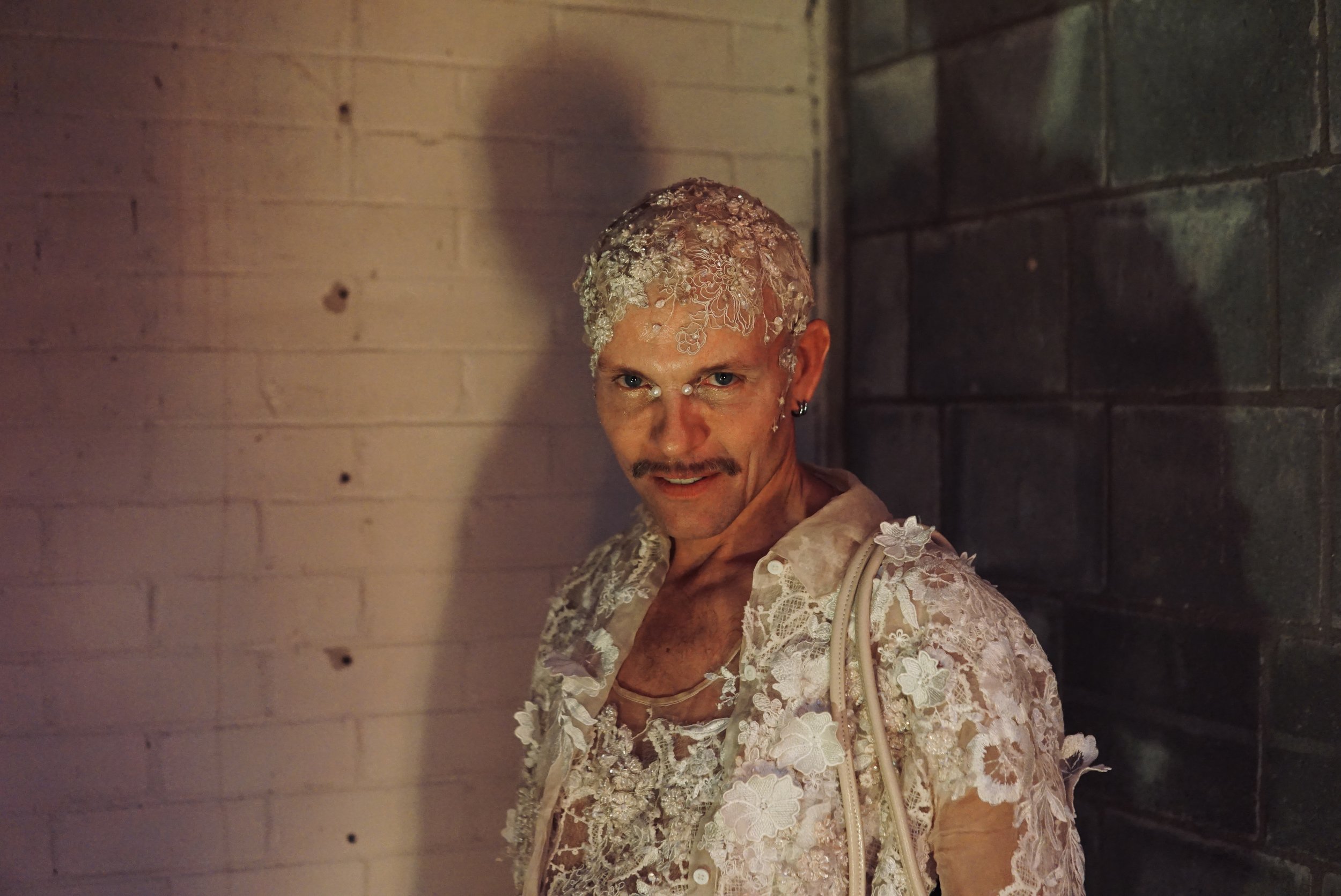 A person with short hair and a mustache, wearing a lace outfit and lace head covering, standing against a mixed brick and concrete wall at night.