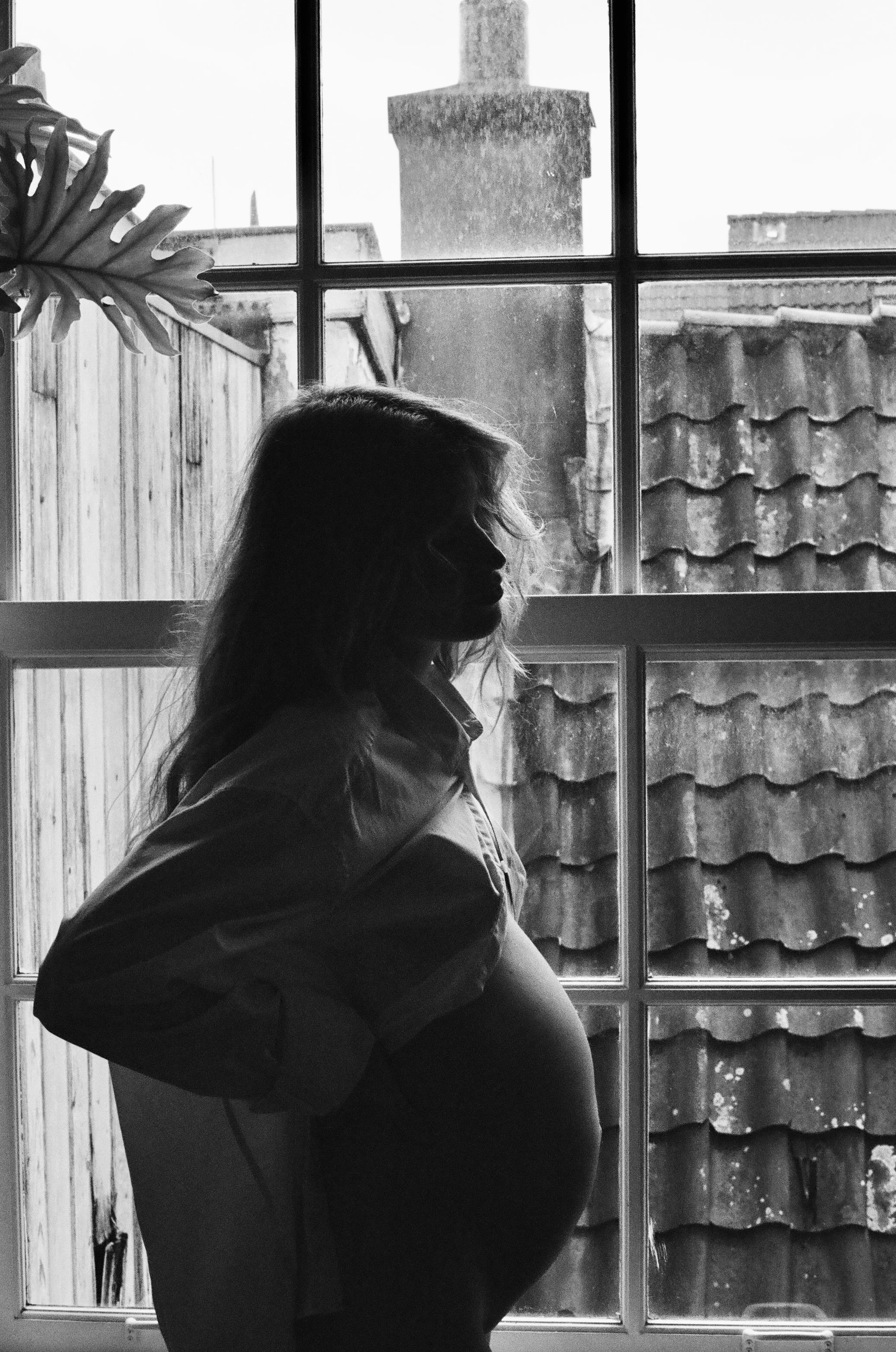 Black and white photo of a pregnant woman standing indoors in front of a window, with a roof and chimney visible outside.