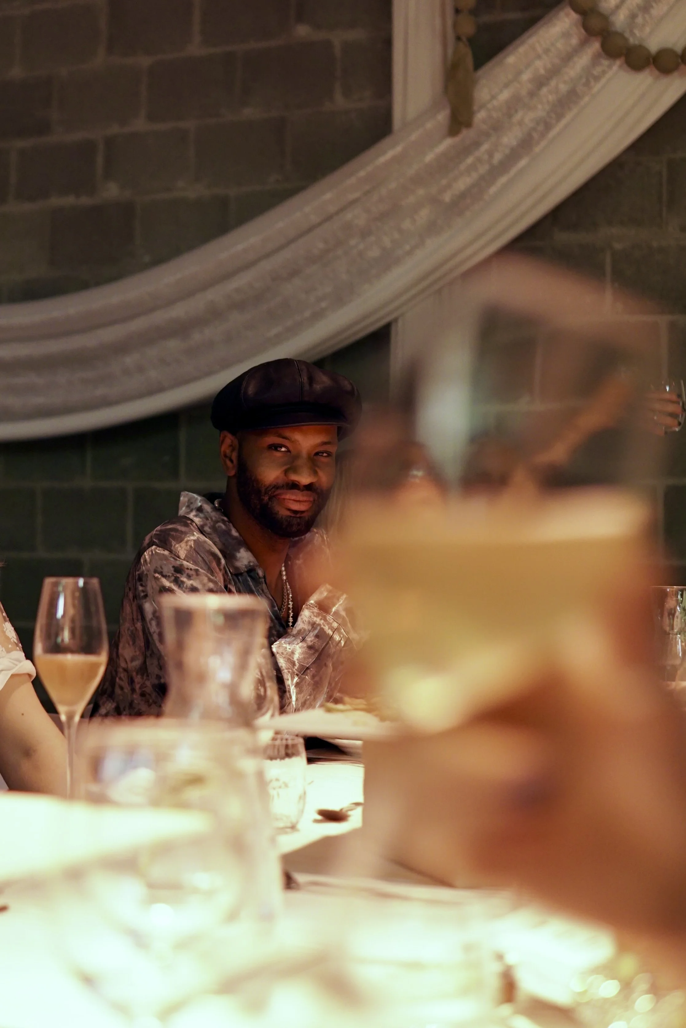 A man wearing a leather hat and patterned shirt sitting at a dinner table with drinks. The background includes a brick wall and decorative drapery.