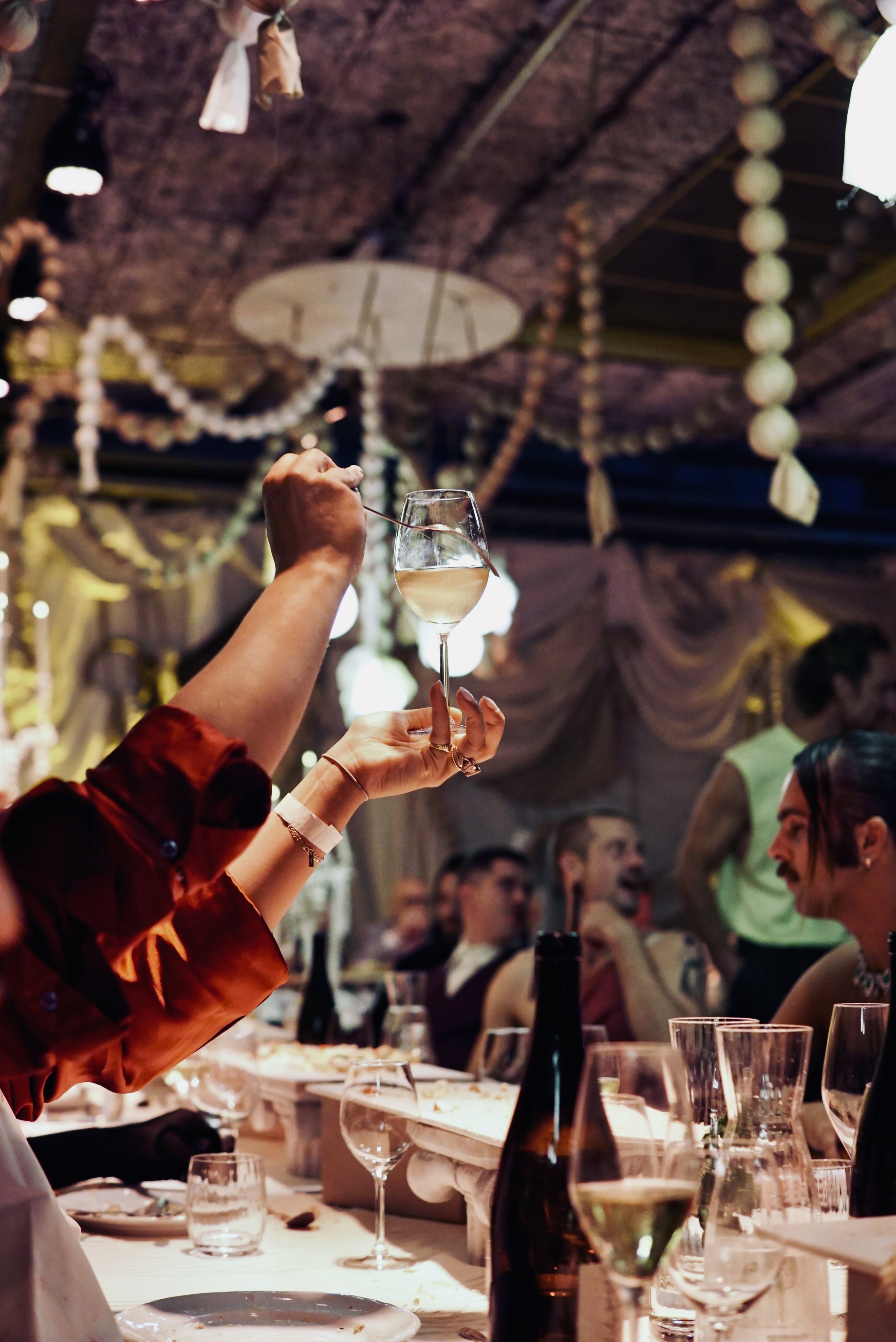 Person holding a glass of white wine at a celebration or party with guests in the background and decorative hanging ornaments.