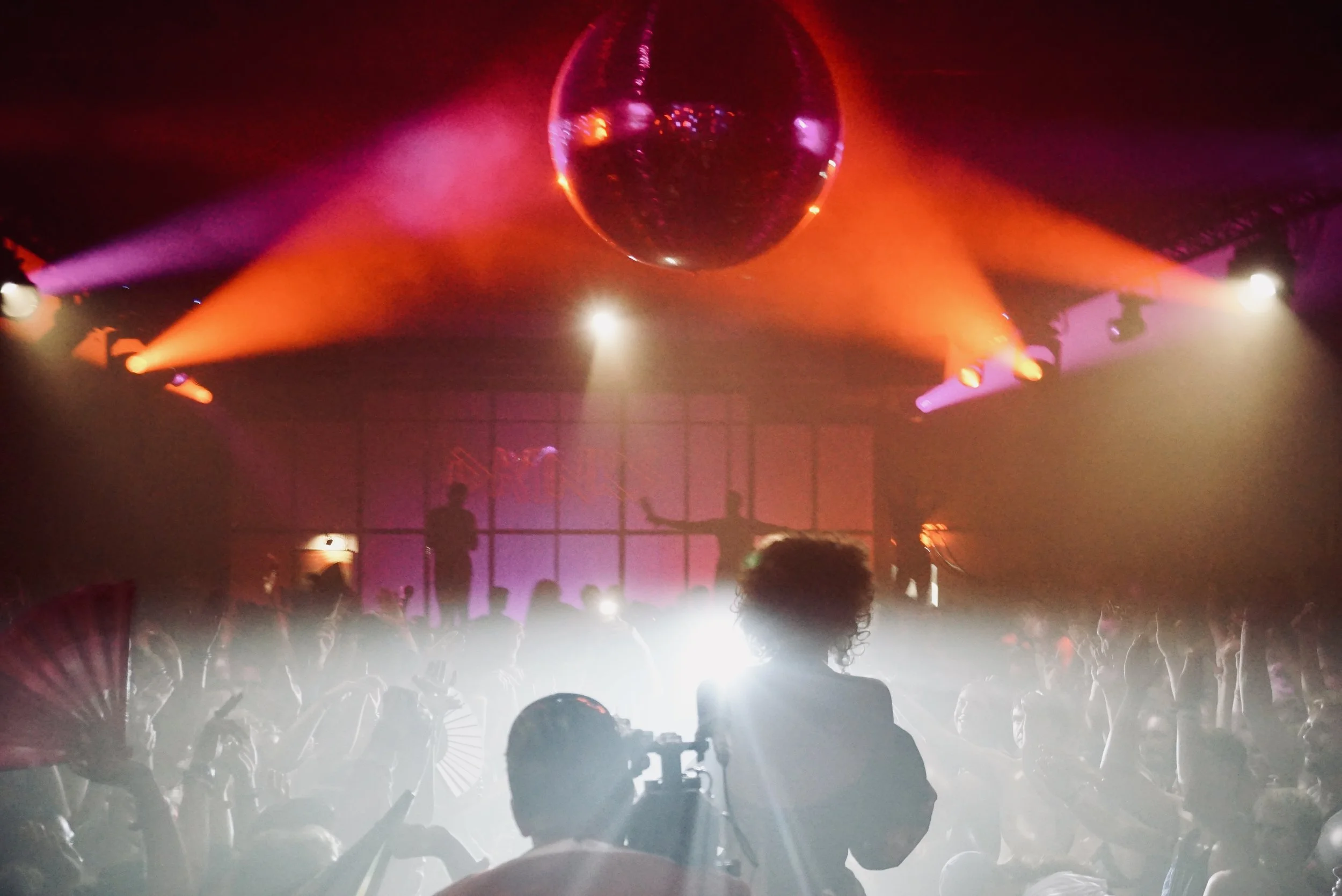 People dancing at a nightclub with colorful lights, a disco ball, and a DJ playing in the background.