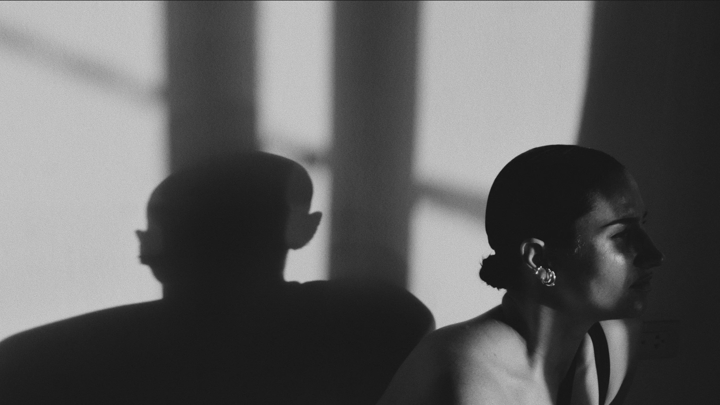 A black and white photo of a woman with short hair and earrings, sitting indoors with shadows cast on the wall behind her.