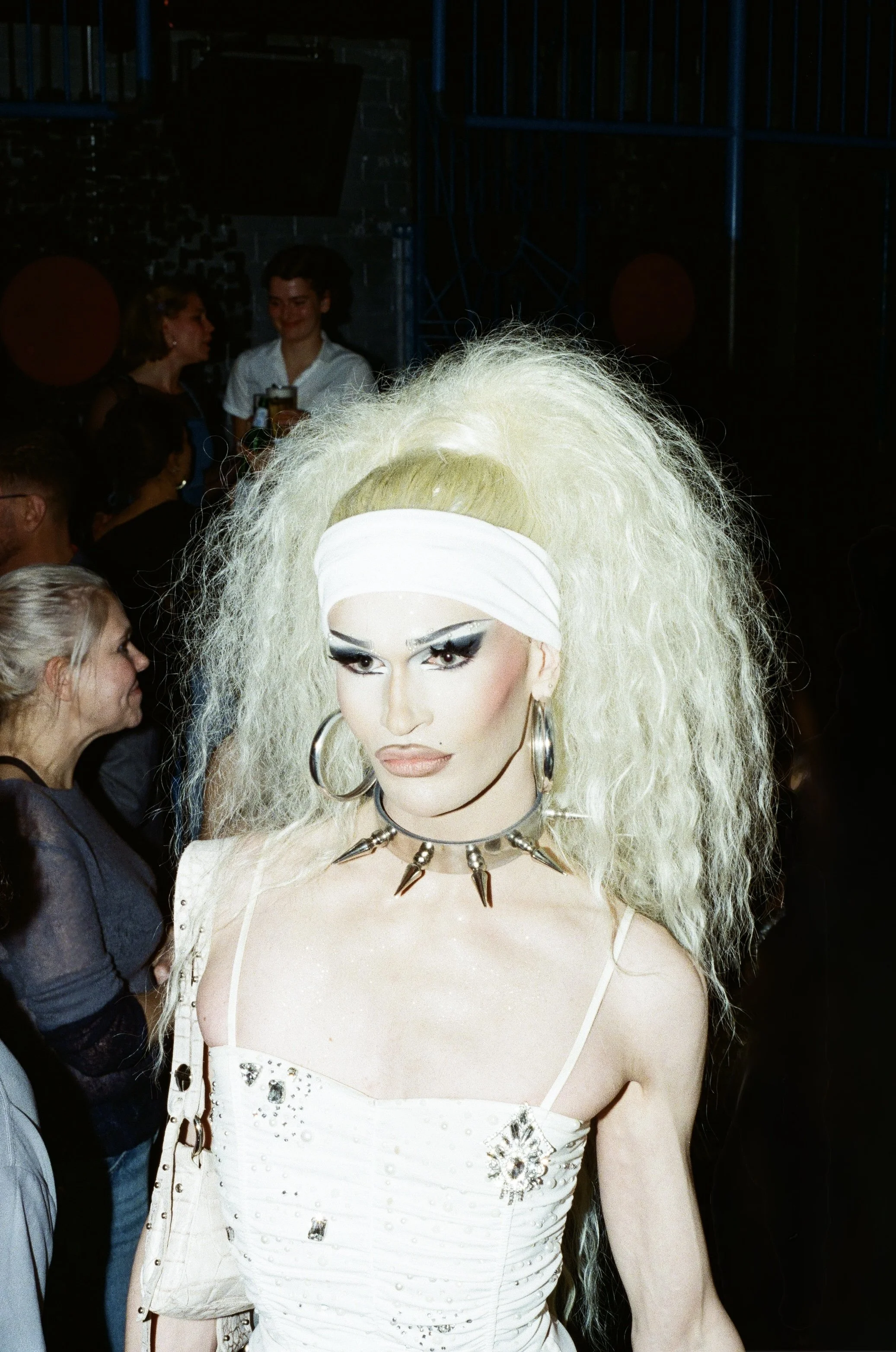 A person with platinum blonde, crimped hair and dramatic makeup in a black and white style, wearing a white headband, large hoop earrings, a spiked choker, and a white dress with embellishments, standing among a crowd in a dimly lit setting.