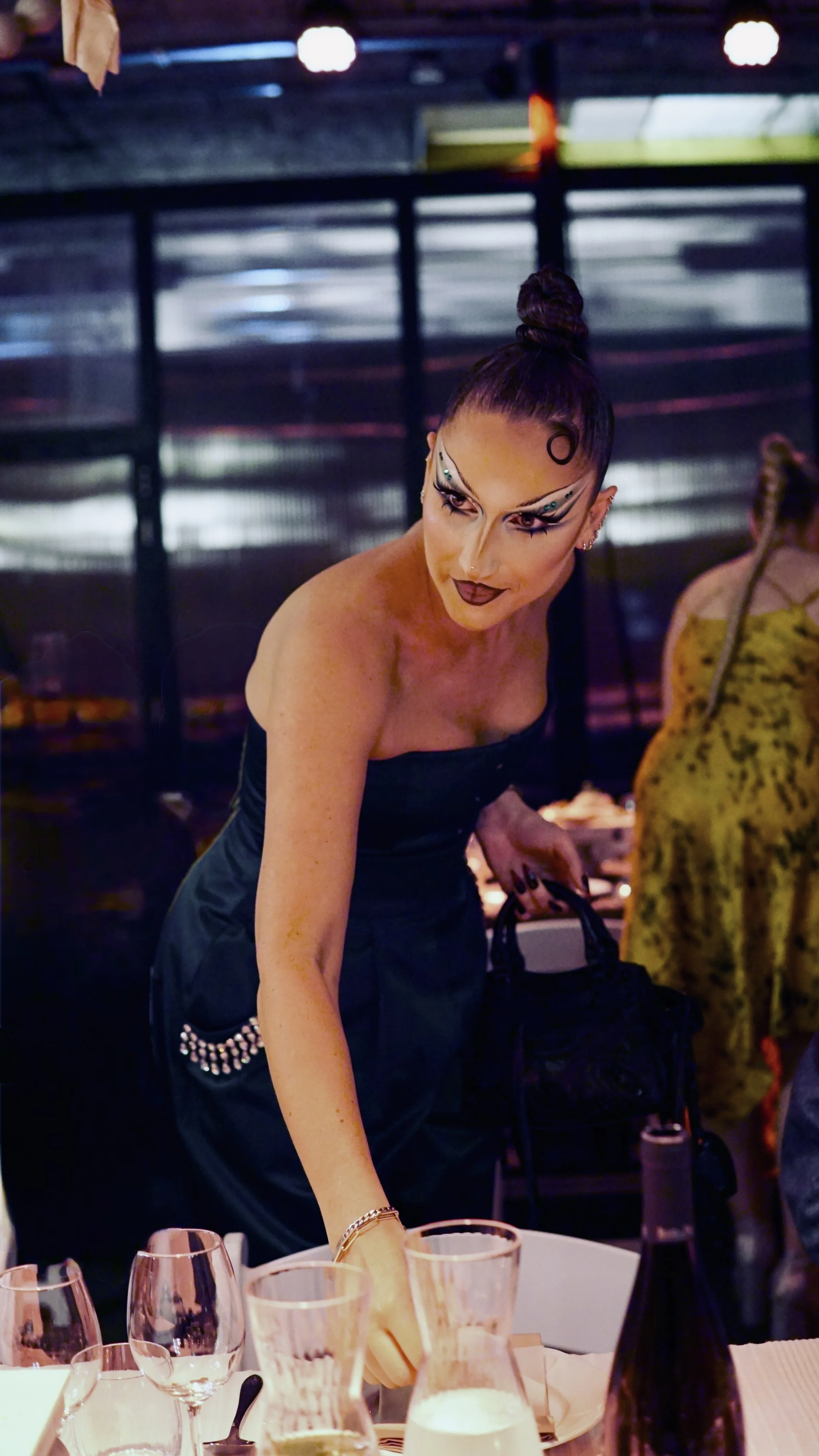 A woman with elaborate makeup and a topknot hairstyle is reaching across a table in a dimly lit setting, possibly a restaurant or event space.