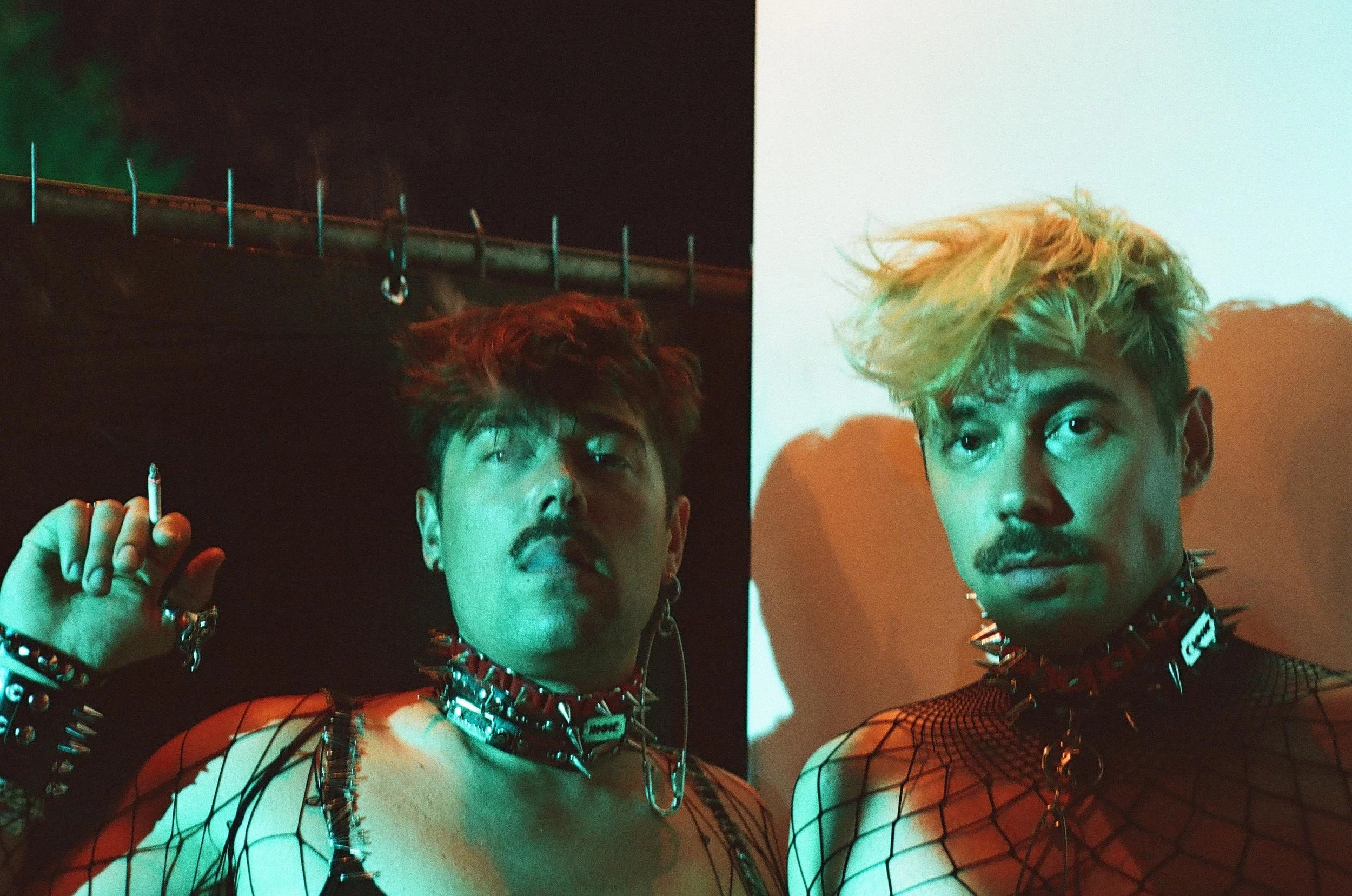 Two men with punk fashion styles pose in a dimly lit photo. The man on the left has dark hair, a mustache, spiked choker, and mesh top, holding a cigarette. The man on the right has bleached hair, facial hair, and wears a similar spiked choker and mesh top, with a serious expression.
