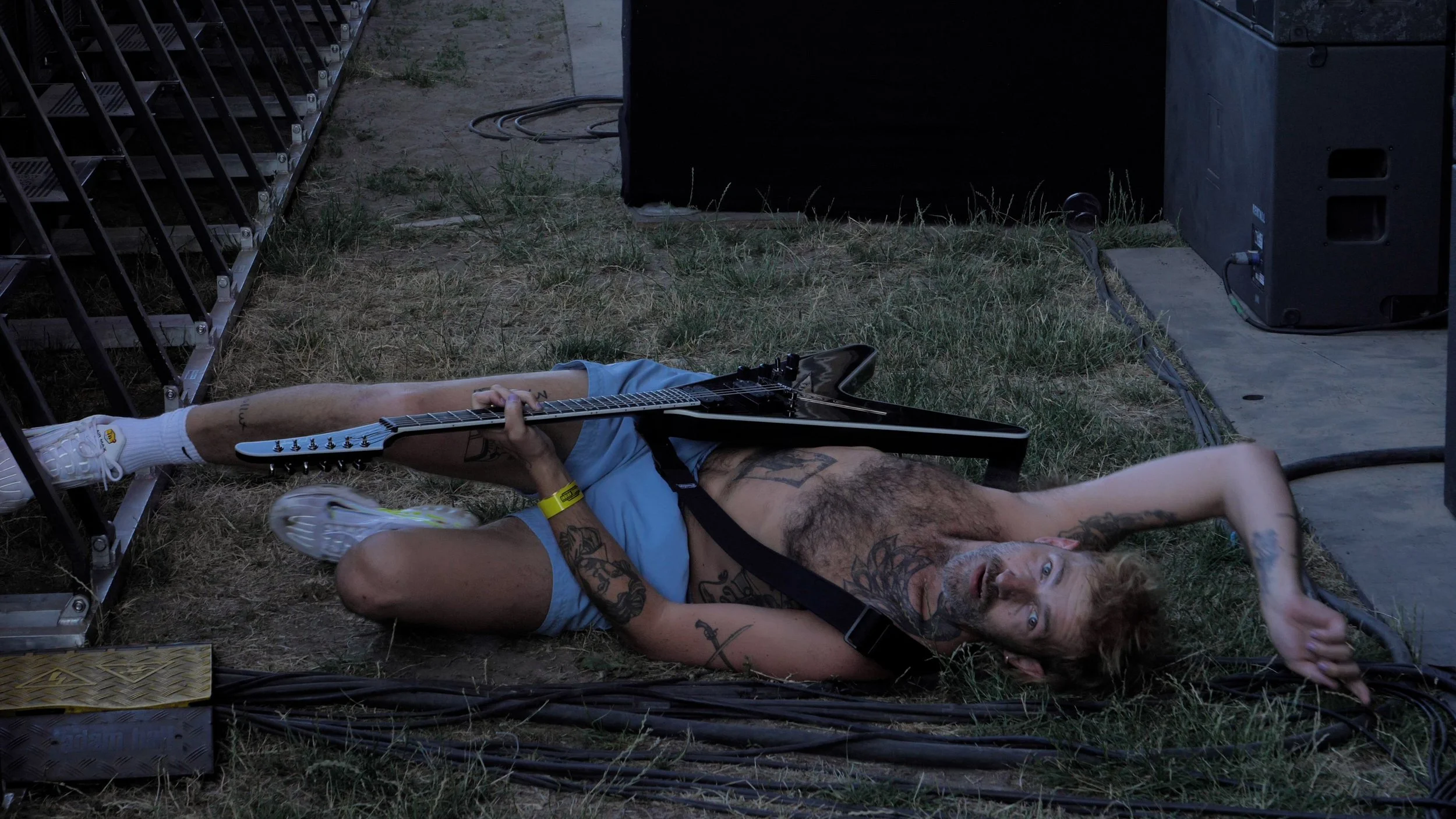 A man lying on the ground at a music event, holding a black electric guitar, with tattoos on his arms and chest, wearing a blue tank top, white sneakers, and a yellow wristband, amidst stage equipment and grass.