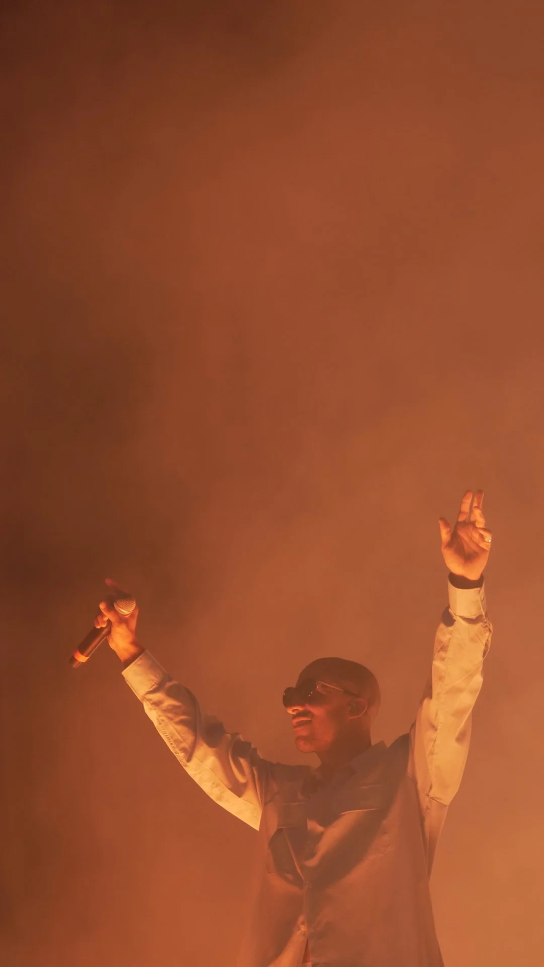 A man wearing sunglasses and a jacket is holding a microphone in one hand and making a peace sign with the other, standing in front of orange-colored fog or smoke.