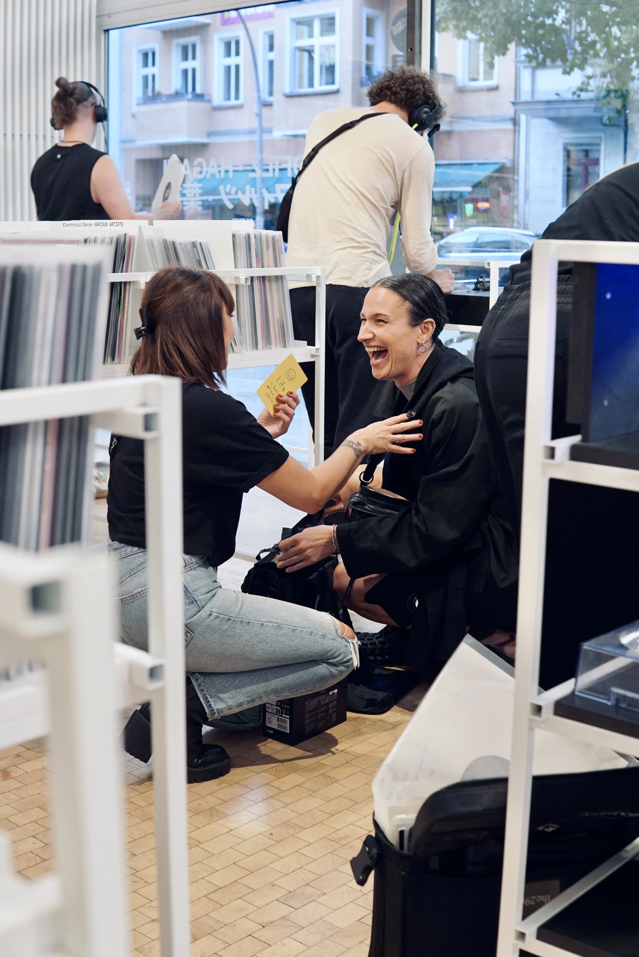 Two women are sitting on the floor of a record store, laughing and smiling at each other. One woman holds a yellow card with a smiley face on it. Behind them, a person with curly hair and wearing a white shirt with headphones is browsing records. The store has large windows, shelves of records, and a view of buildings and trees outside.