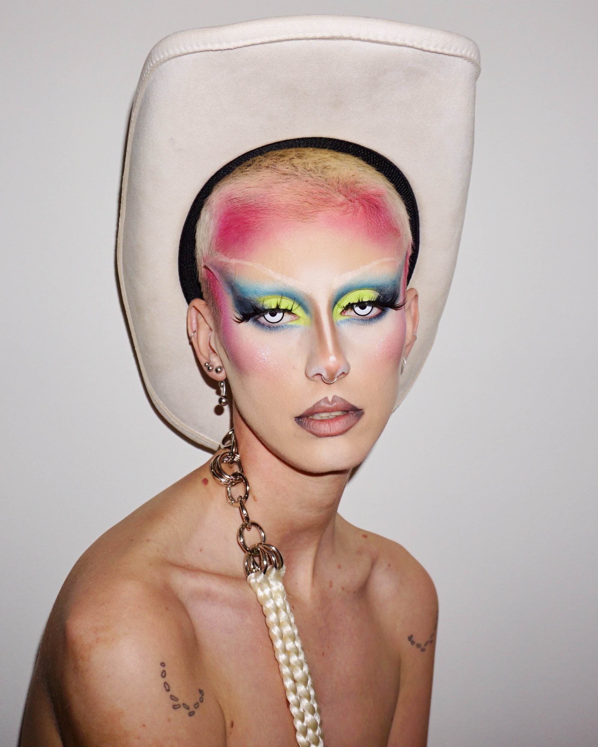 Person with colorful makeup, blond hair with pink dye, large white hat, multiple earrings, and a pearl necklace.