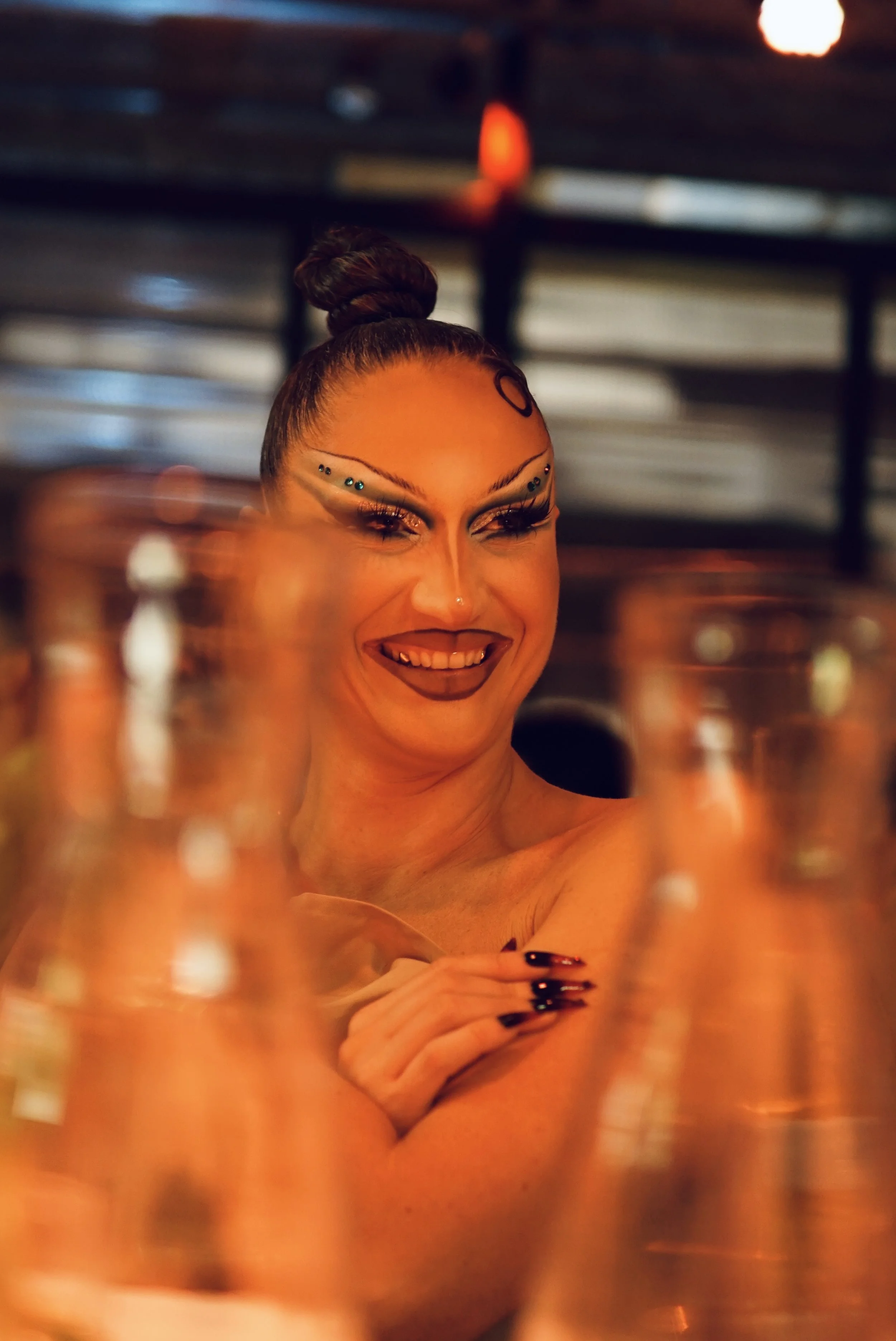 A woman smiling with elaborate makeup and jewelry, surrounded by glass bottles in a dimly lit setting.