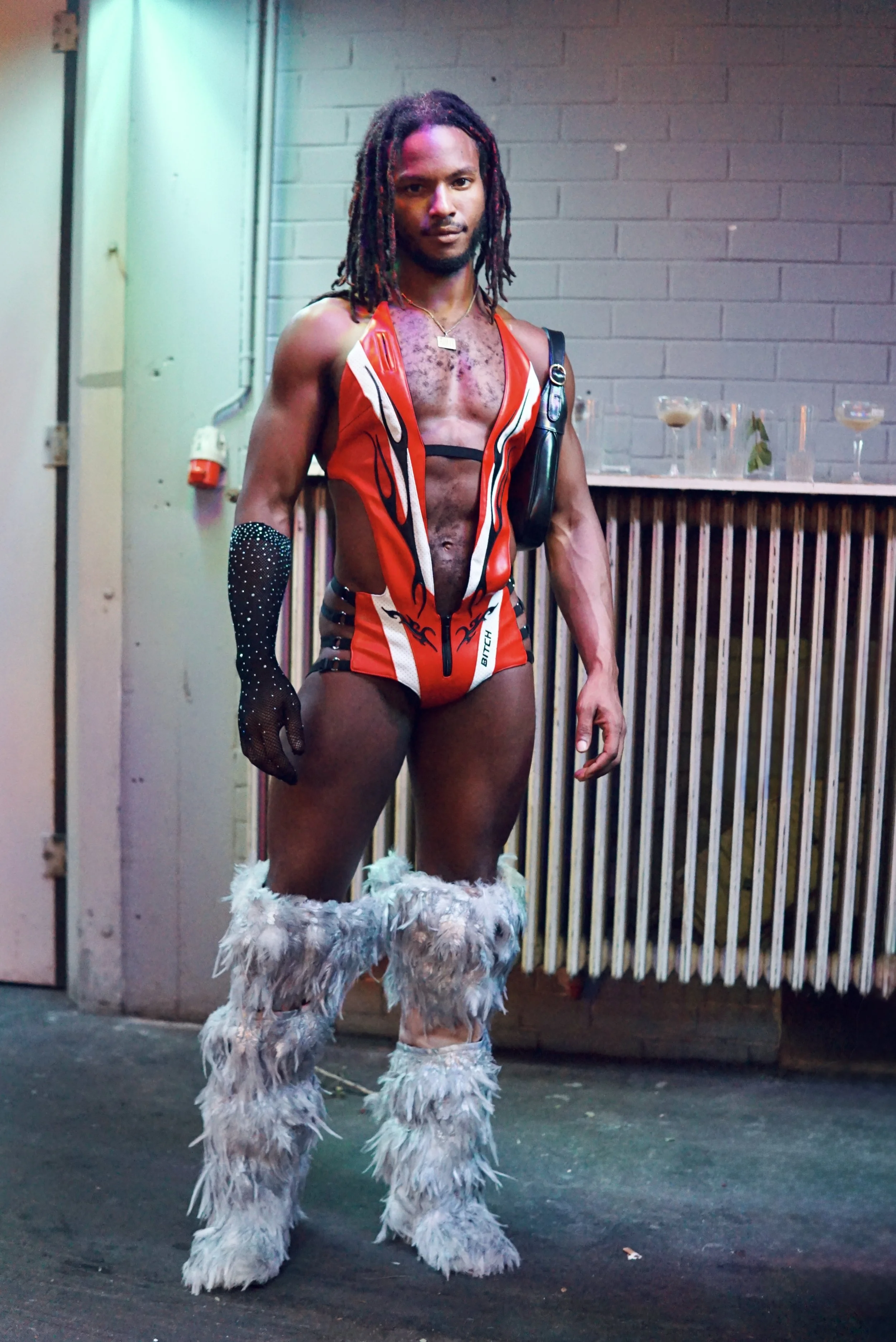 A man with dreadlocks wearing a red and white sleeveless vest, furry white boots, and a black glove with rhinestones stands in front of a brick wall.