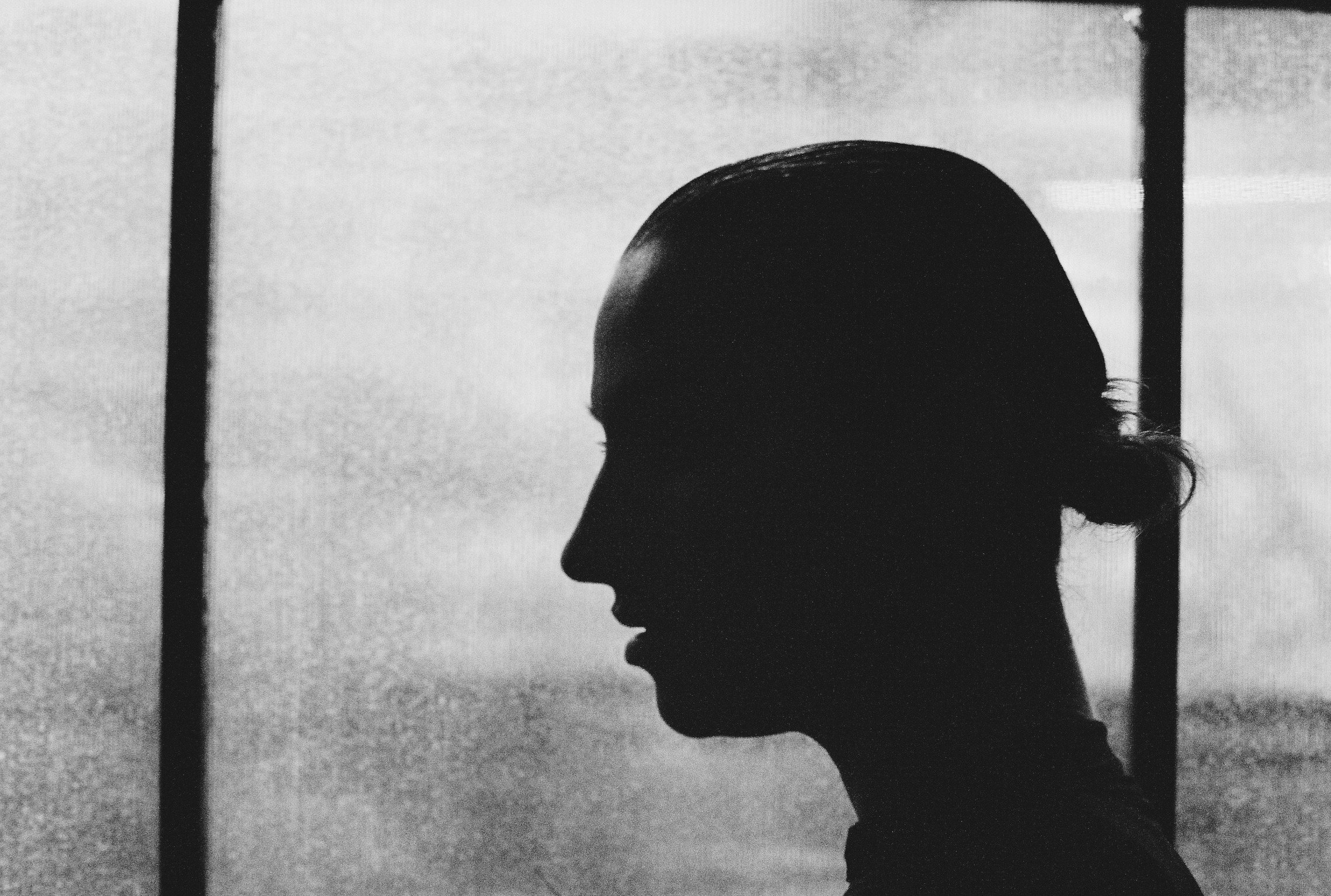 Black and white silhouette of a woman with a bun hairstyle, facing left, with a background of window frames and cloudy sky.