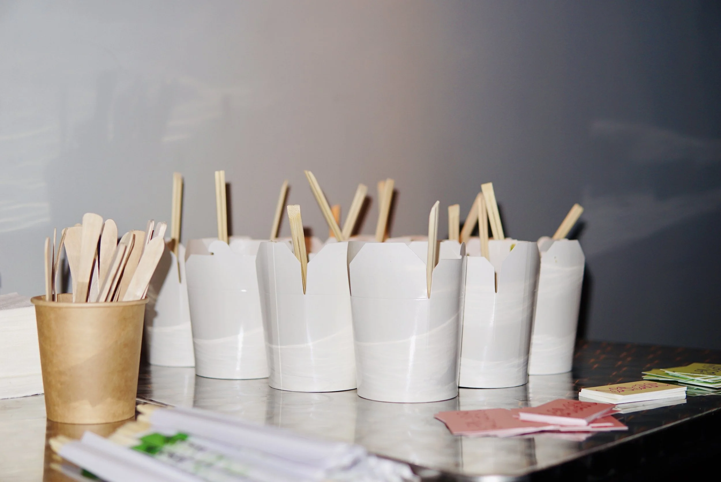 Multiple white disposable cups filled with wooden stir sticks on a table, with a small paper cup holding additional stir sticks nearby, and scattered paper sheets and cards in the foreground.