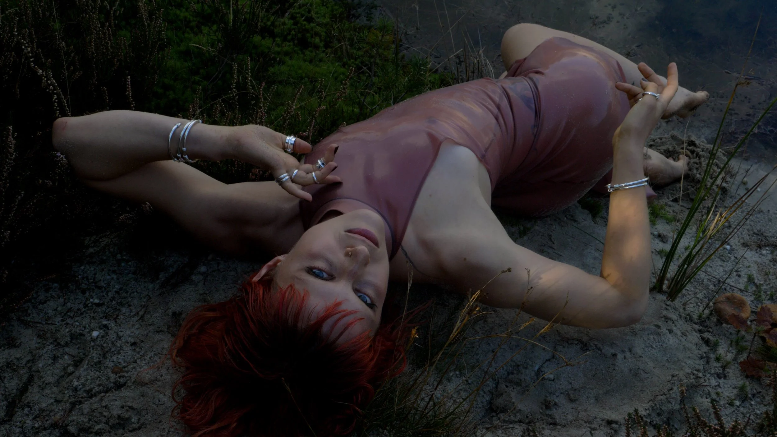 Woman with red hair lying on the ground near a body of water, wearing a sleeveless top and multiple silver rings and bracelets, looking at the camera.