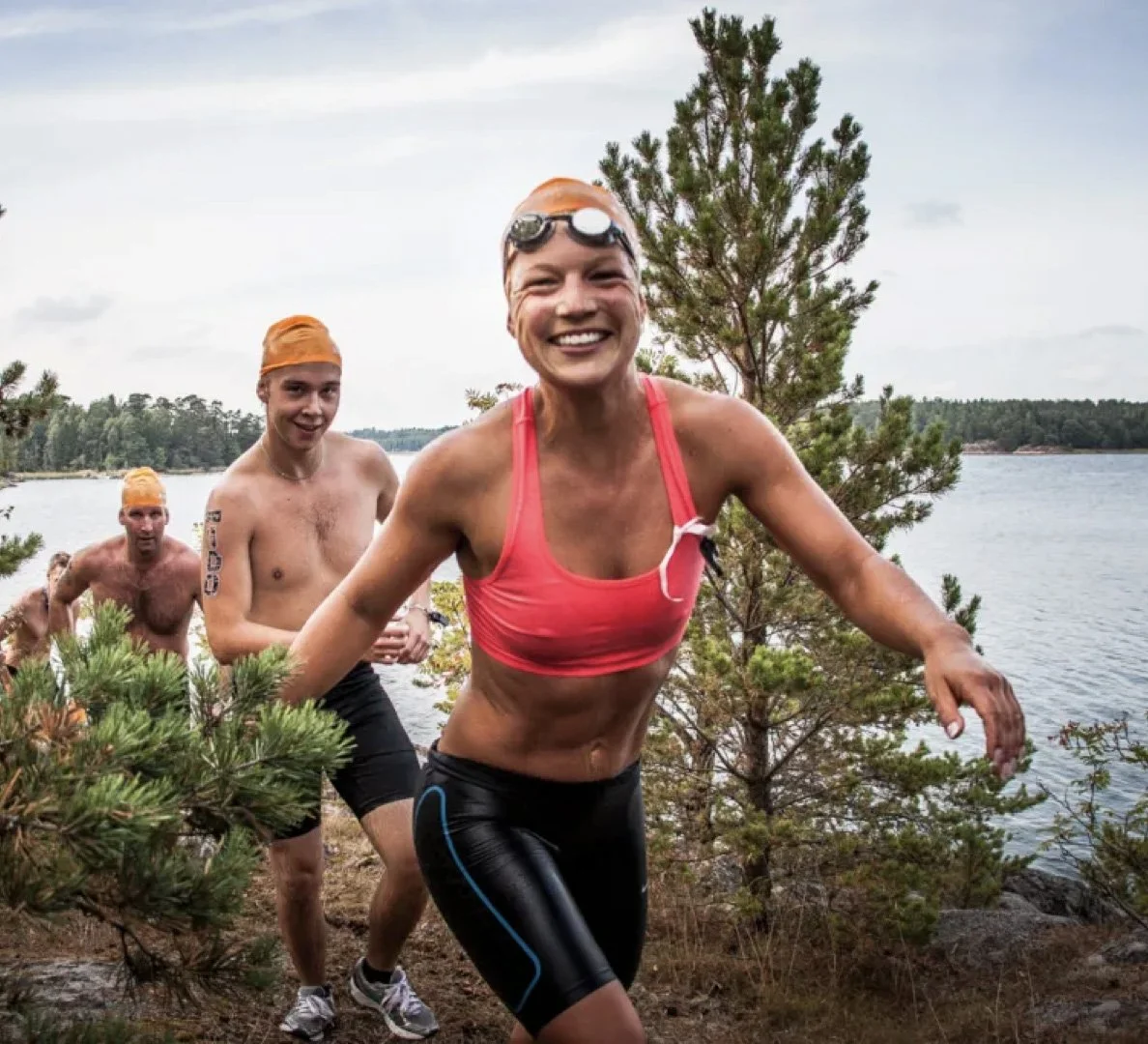 Grupp människor i triathlon, inklusive en kvinna i rosa sporttop och svarta cykelbyxor, som springer längs stranden vid en sjö, omgivna av träd och natur under en molnig dag.