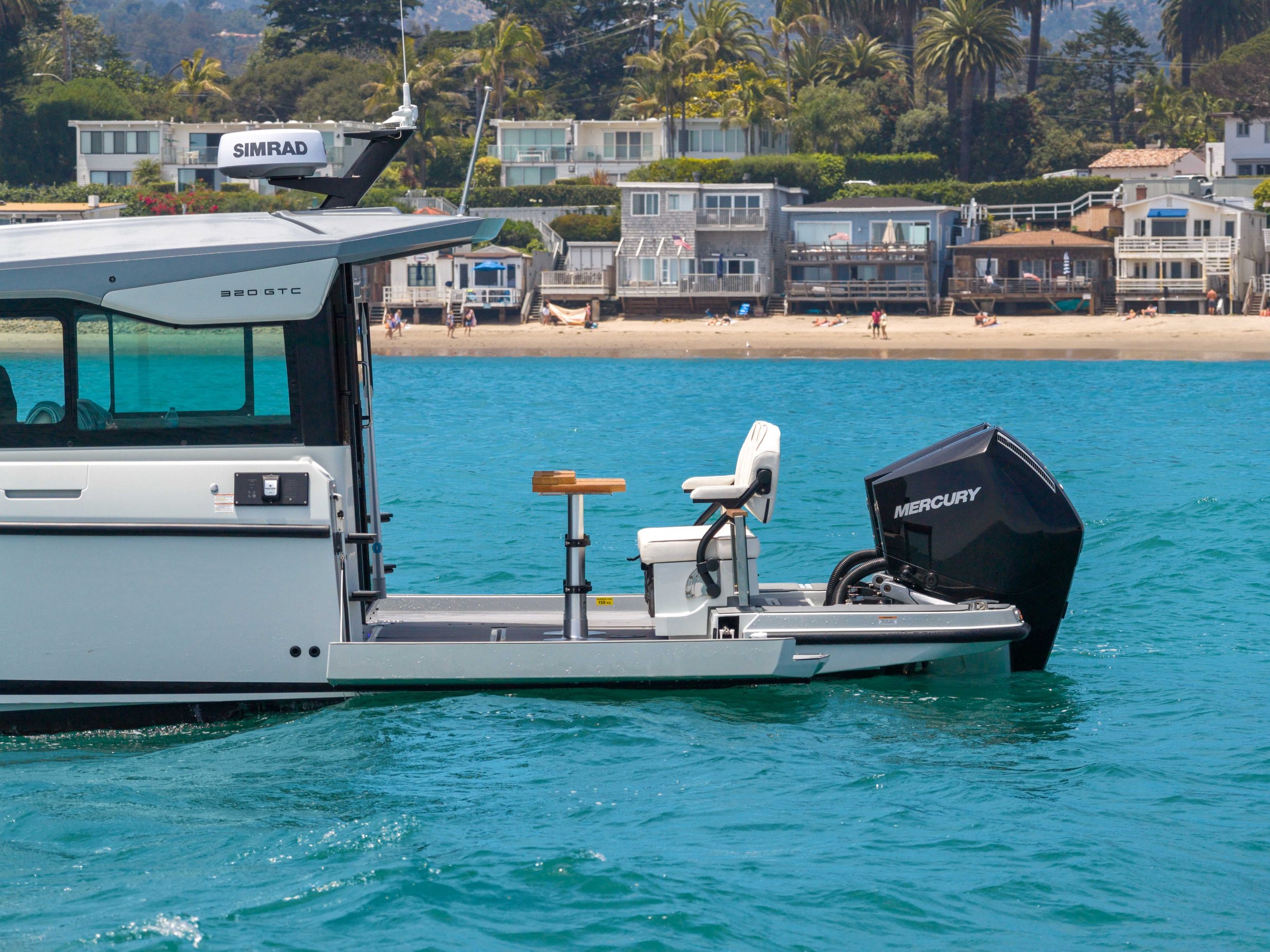 Close-up of a boat with an outboard motor and navigation equipment, floating on turquoise water near houses and trees on a coastal shoreline.