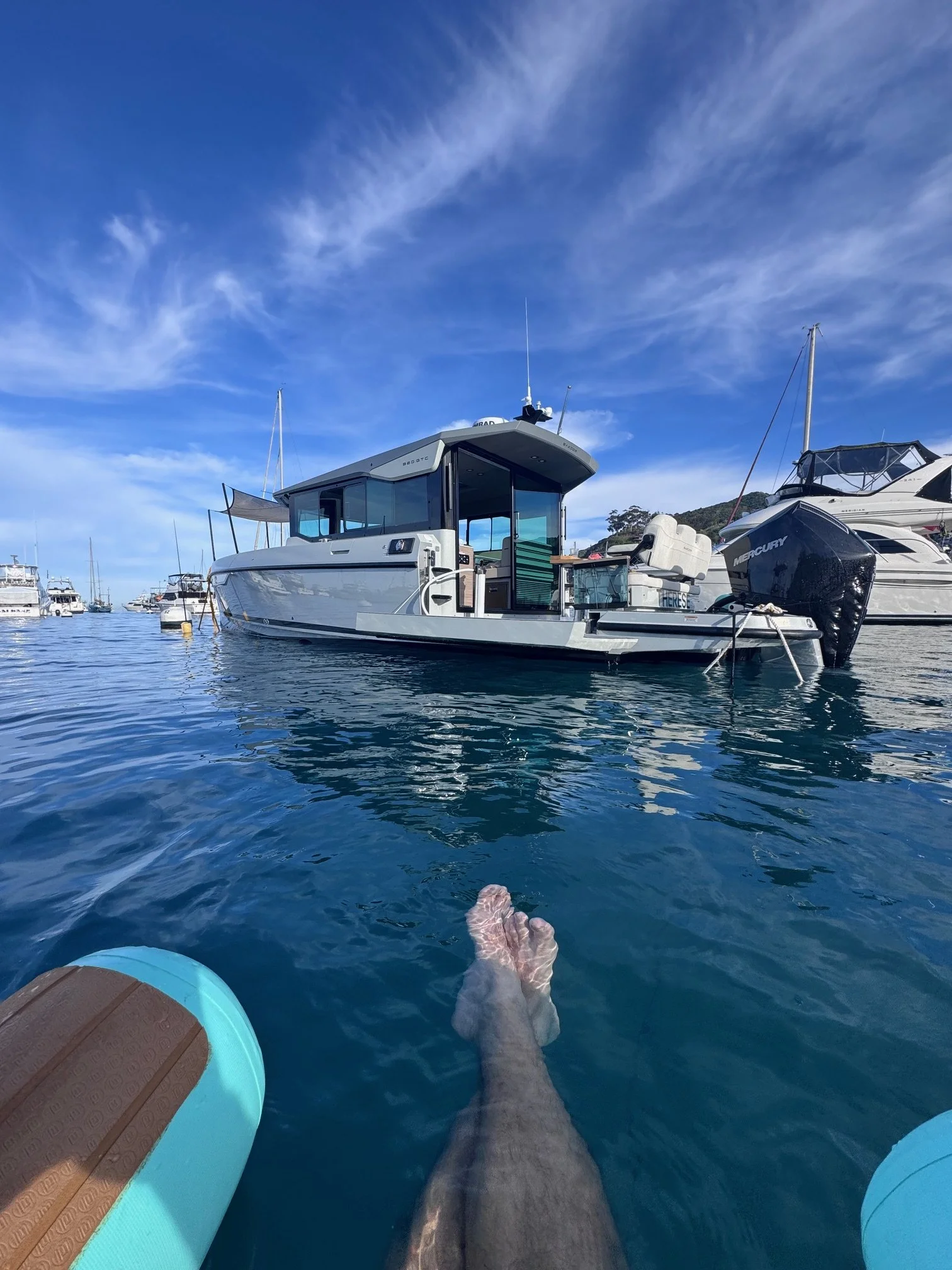 Person floating in a body of water on a blue inflatable tube, with a white boat docked nearby and more boats in the background under a partly cloudy blue sky.
