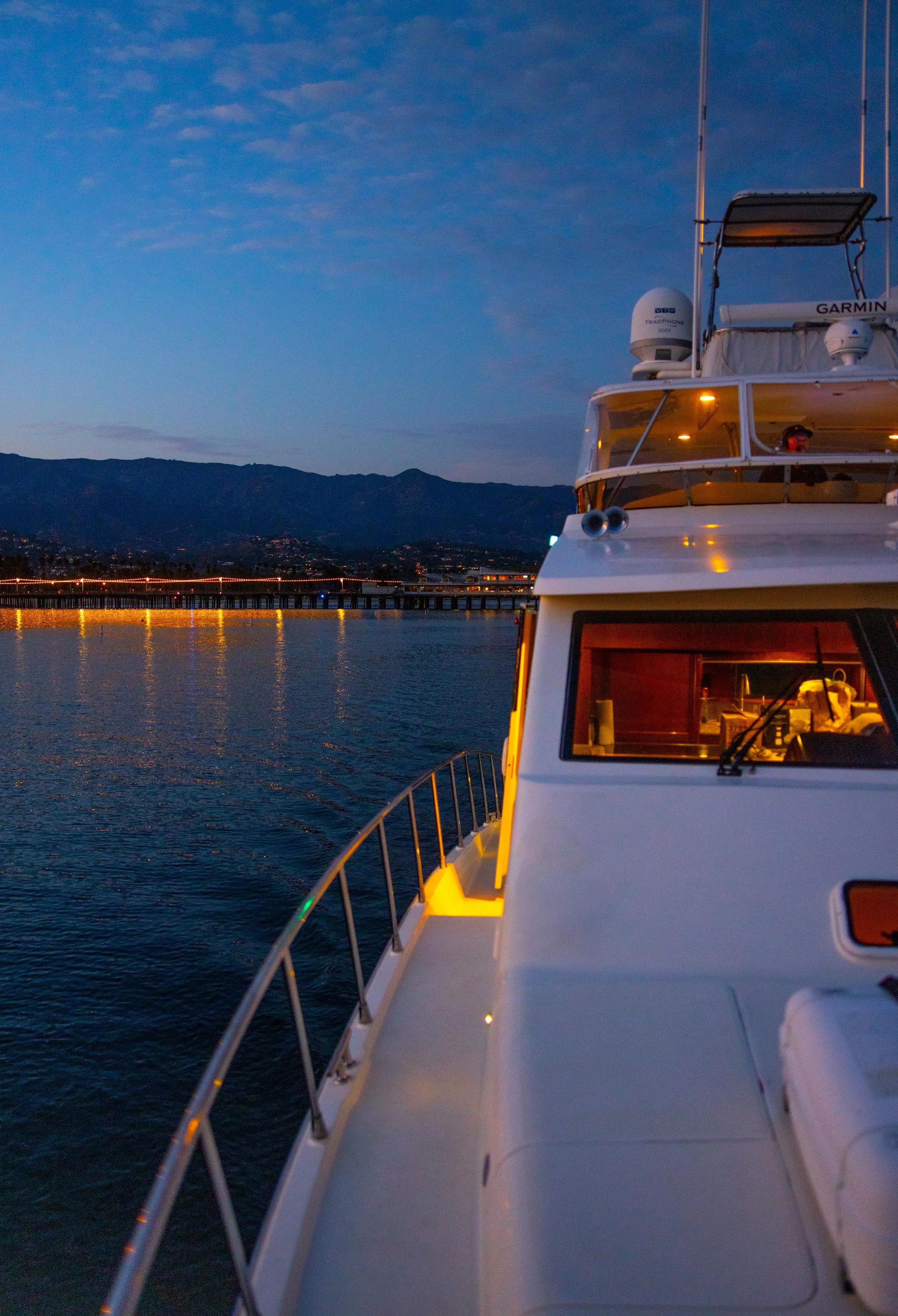 A yacht docked on calm water during twilight, with mountains in the background and city lights on a bridge, and a person wearing headphones inside the yacht.