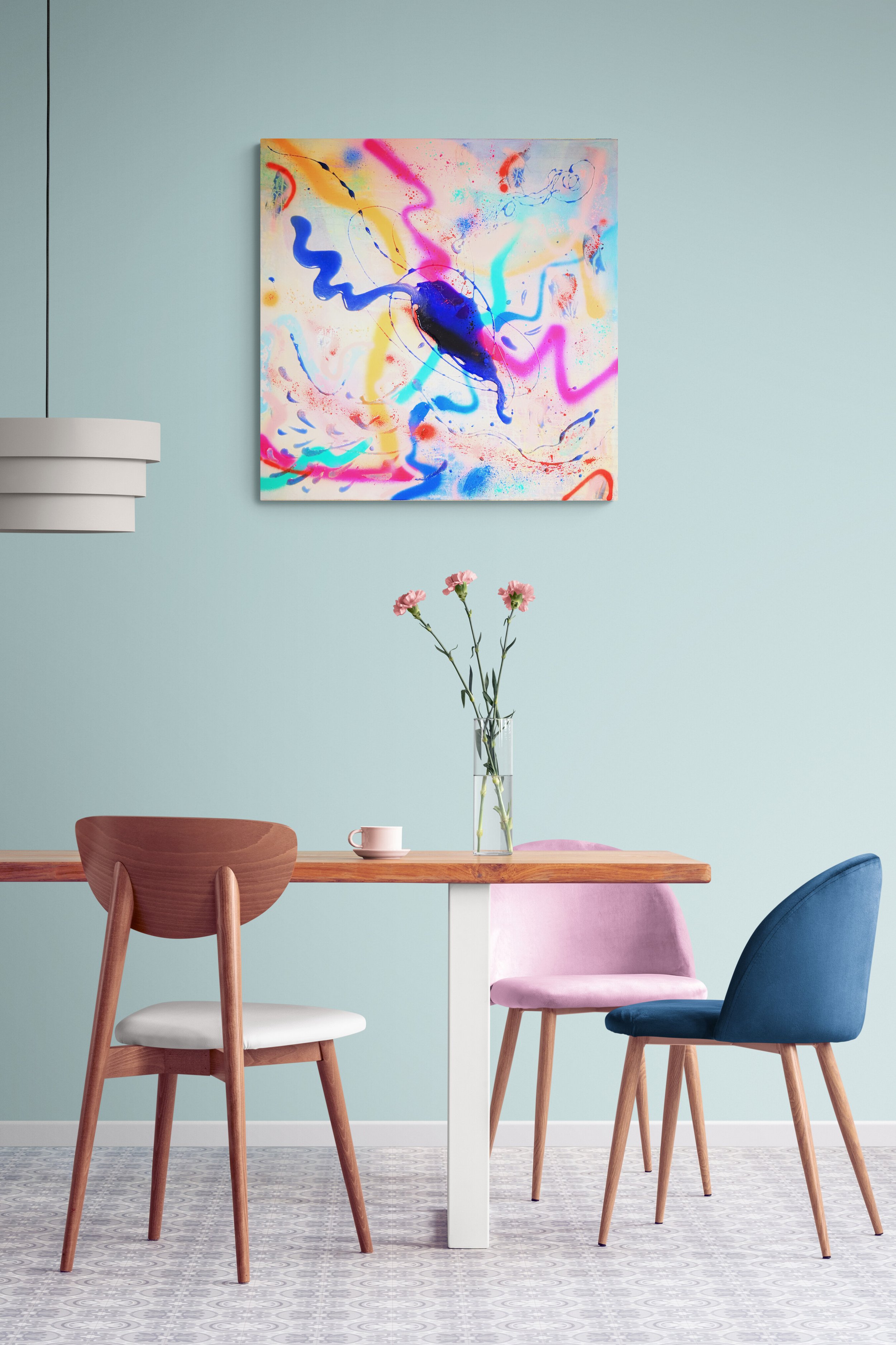 A modern dining room with a wooden table and four colorful chairs in pink, blue, beige, and wood. There is a clear vase with pink flowers on the table. A pastel blue wall features an abstract colorful painting, and a contemporary white light fixture is partially visible on the left. The room has a patterned gray and white tiled floor.