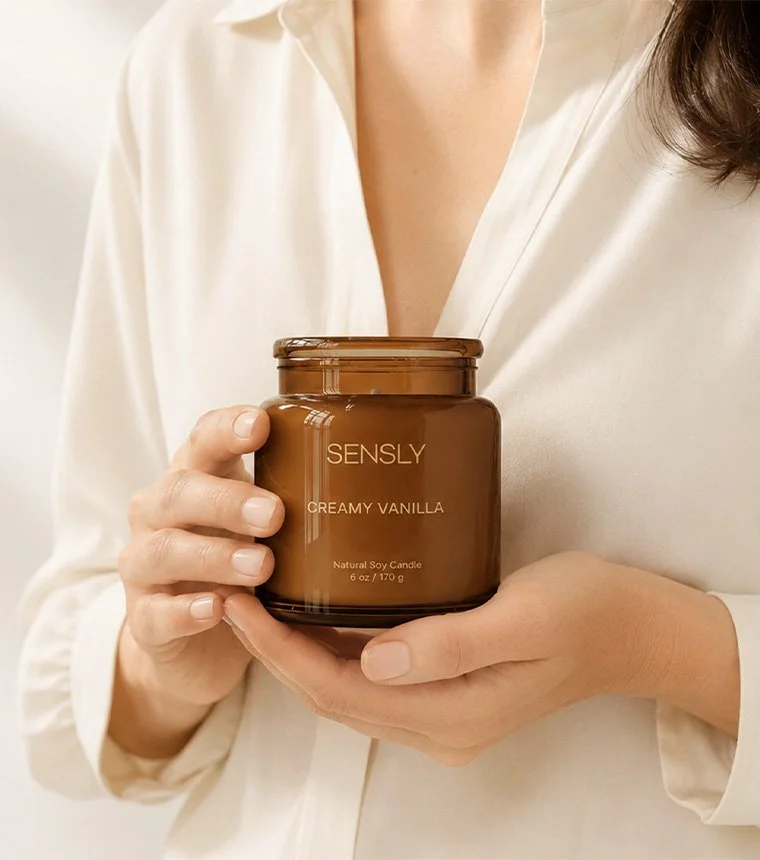 Person holding a brown jar labeled 'Sensly Creamy Vanilla' candle, wearing a satin white blouse.