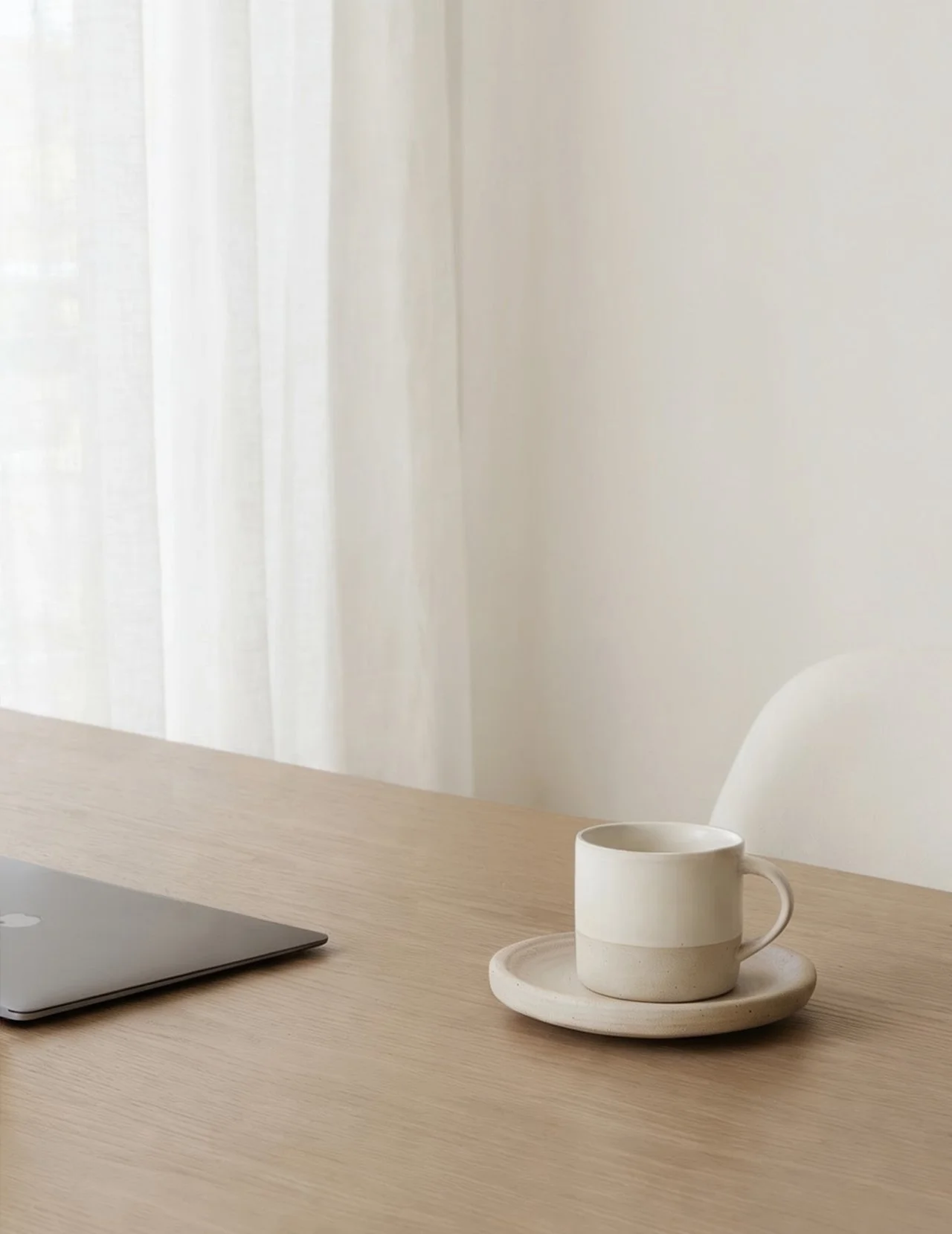 A wooden desk with a closed laptop, a white ceramic mug on a matching plate and saucer, and a white chair in a room with white curtains and walls.